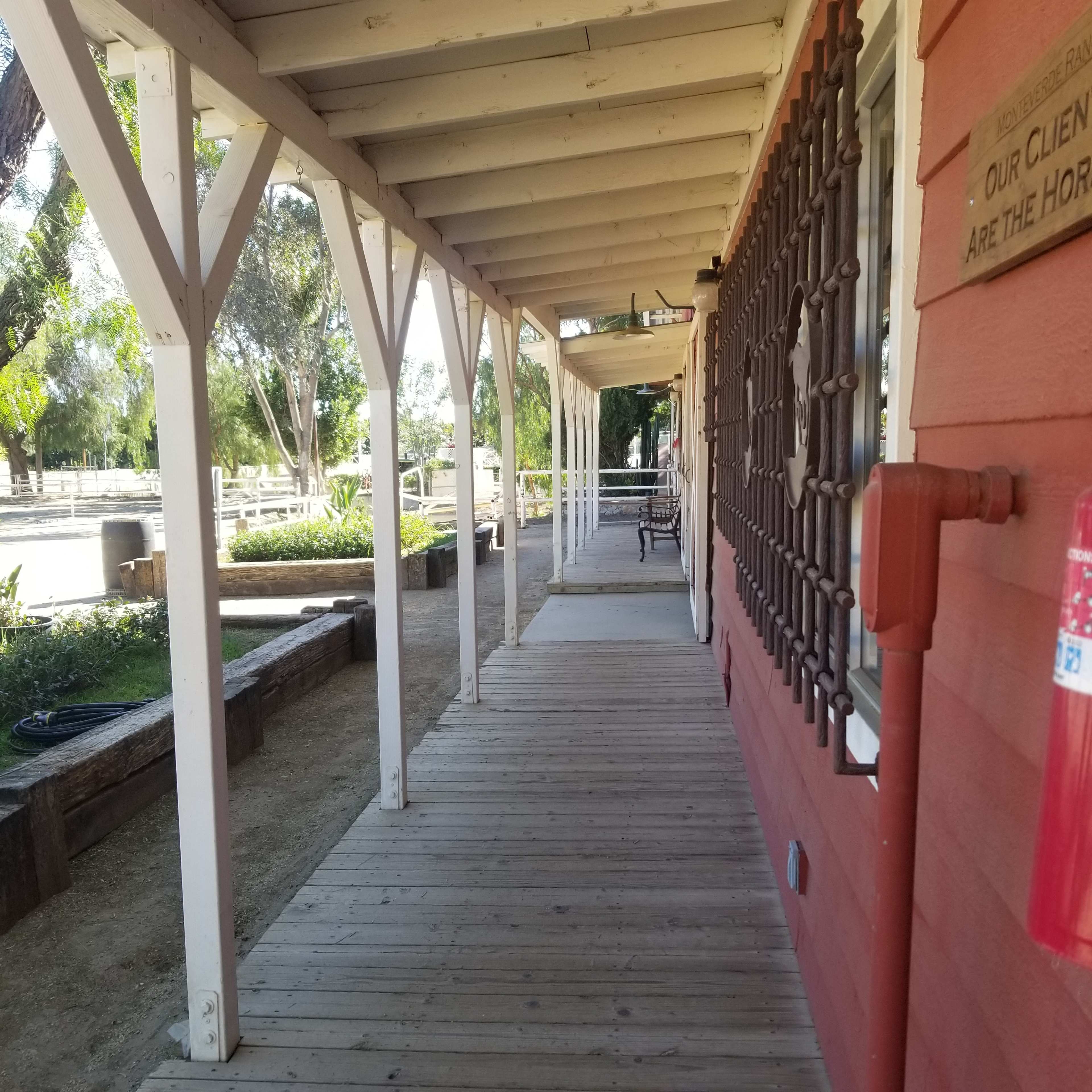 A wooden walkway lined by a building with red siding and a metal grate on a window, leading to an open area with tables and trees in the background.