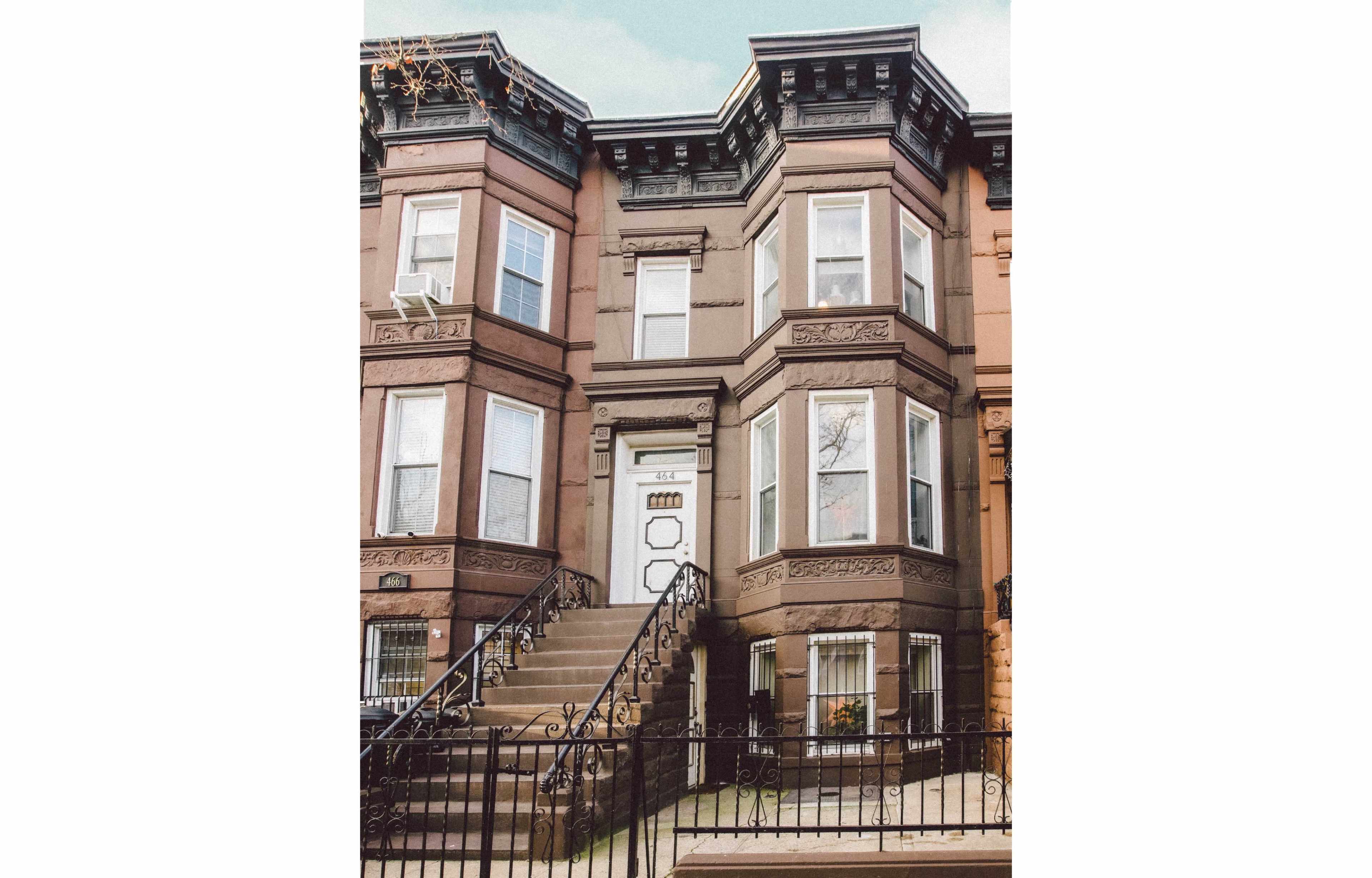 A brownstone building with a set of stairs leading to a door framed by large windows on either side.