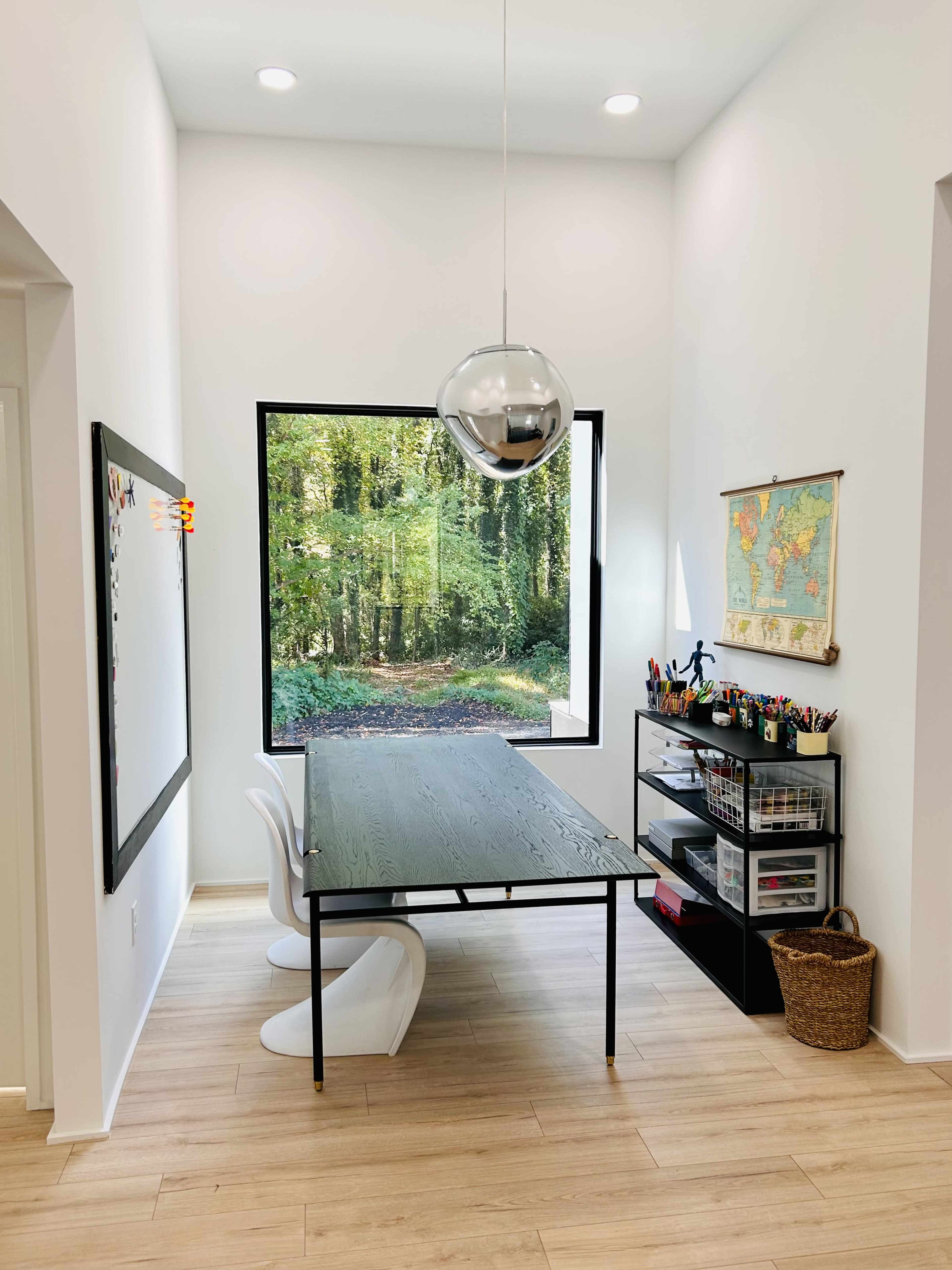 A modern workspace features a black table with white chairs, a large window overlooking a forest, and a wall-mounted map alongside a shelf with art supplies.