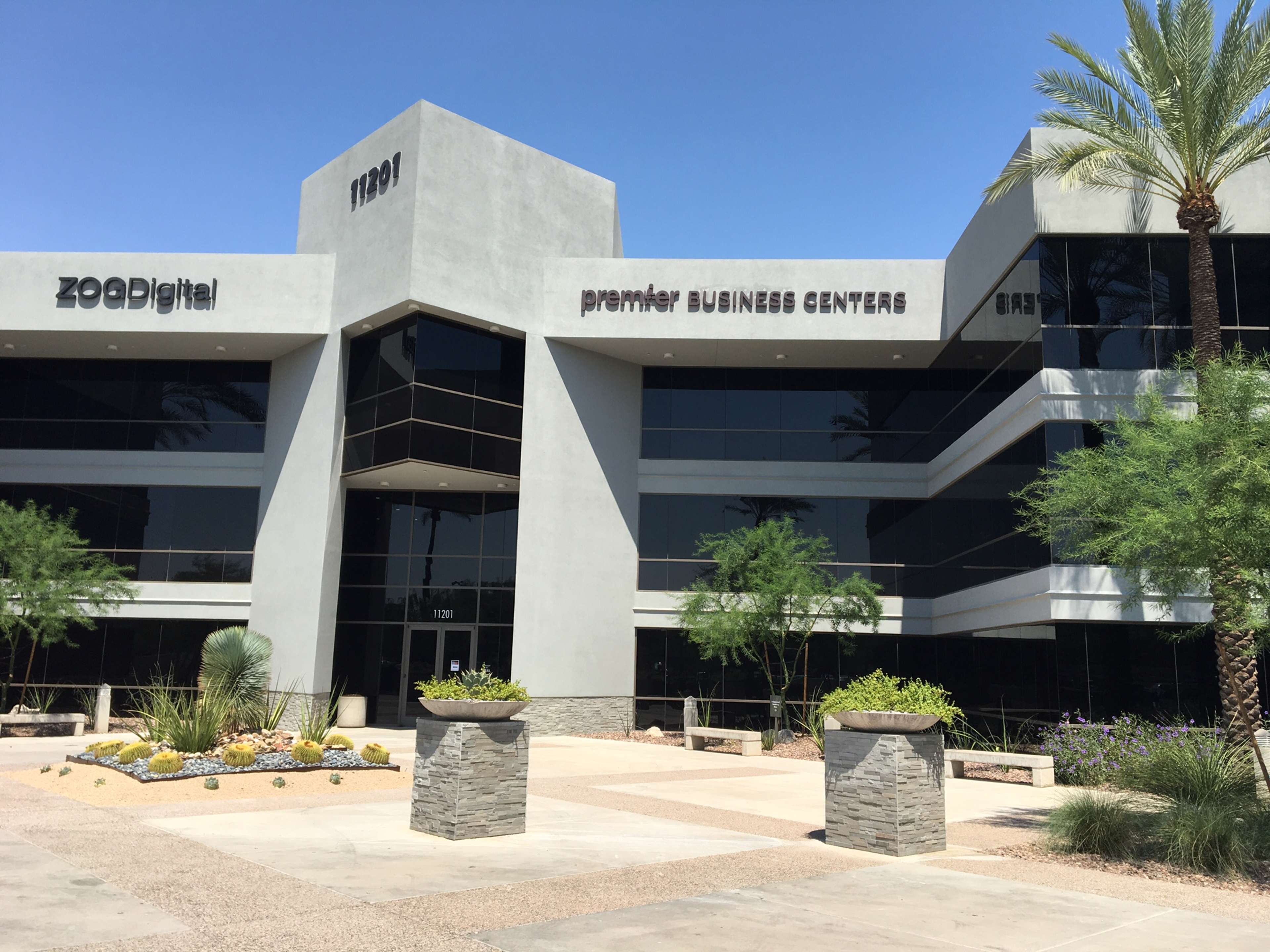 A modern commercial building with a glass facade and landscaped areas, featuring signage for "ZOODigital" and "premier BUSINESS CENTERS."