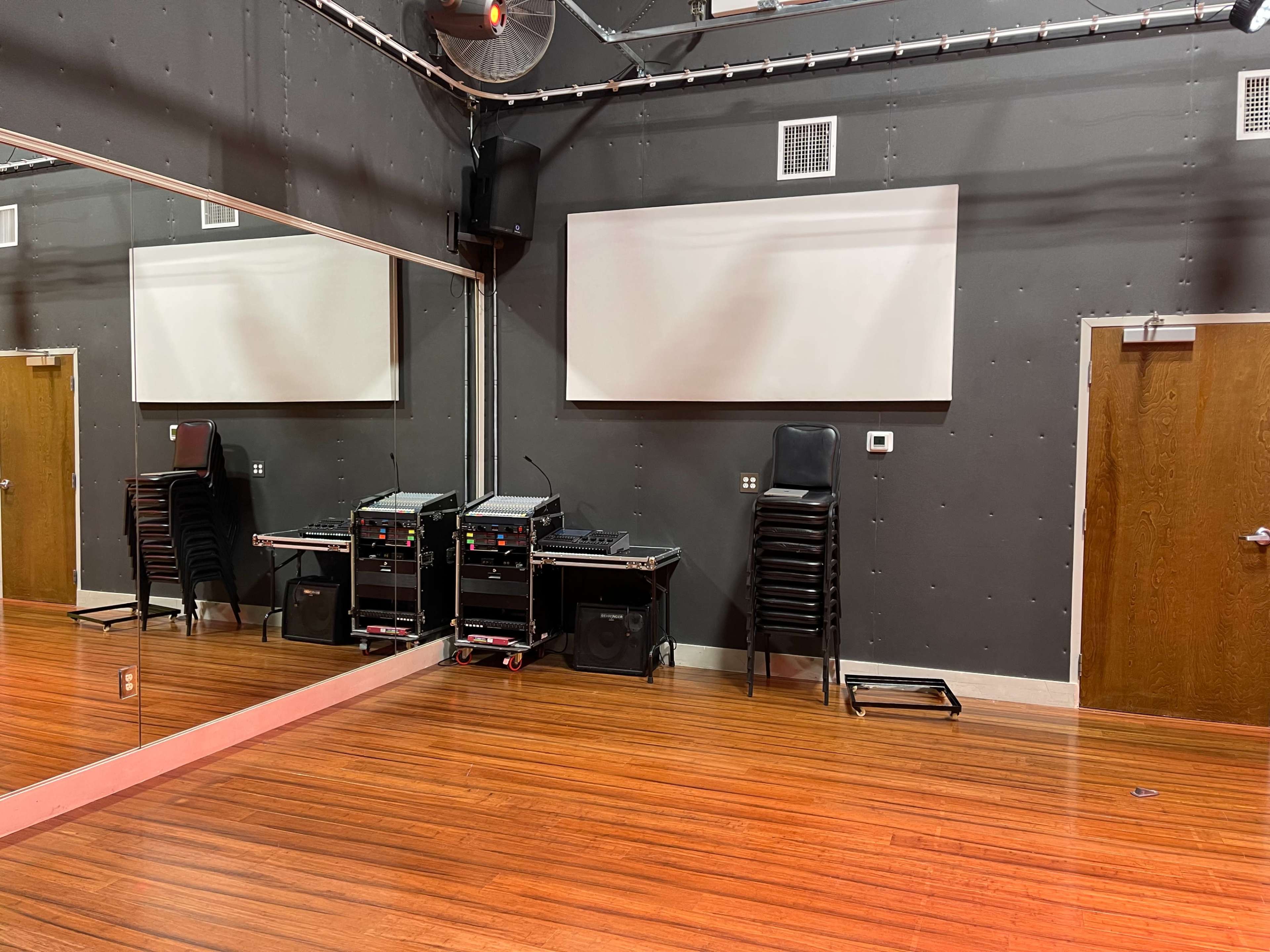 A rehearsal studio with mirrors on one wall, stacked black chairs, and audio equipment in the corner.