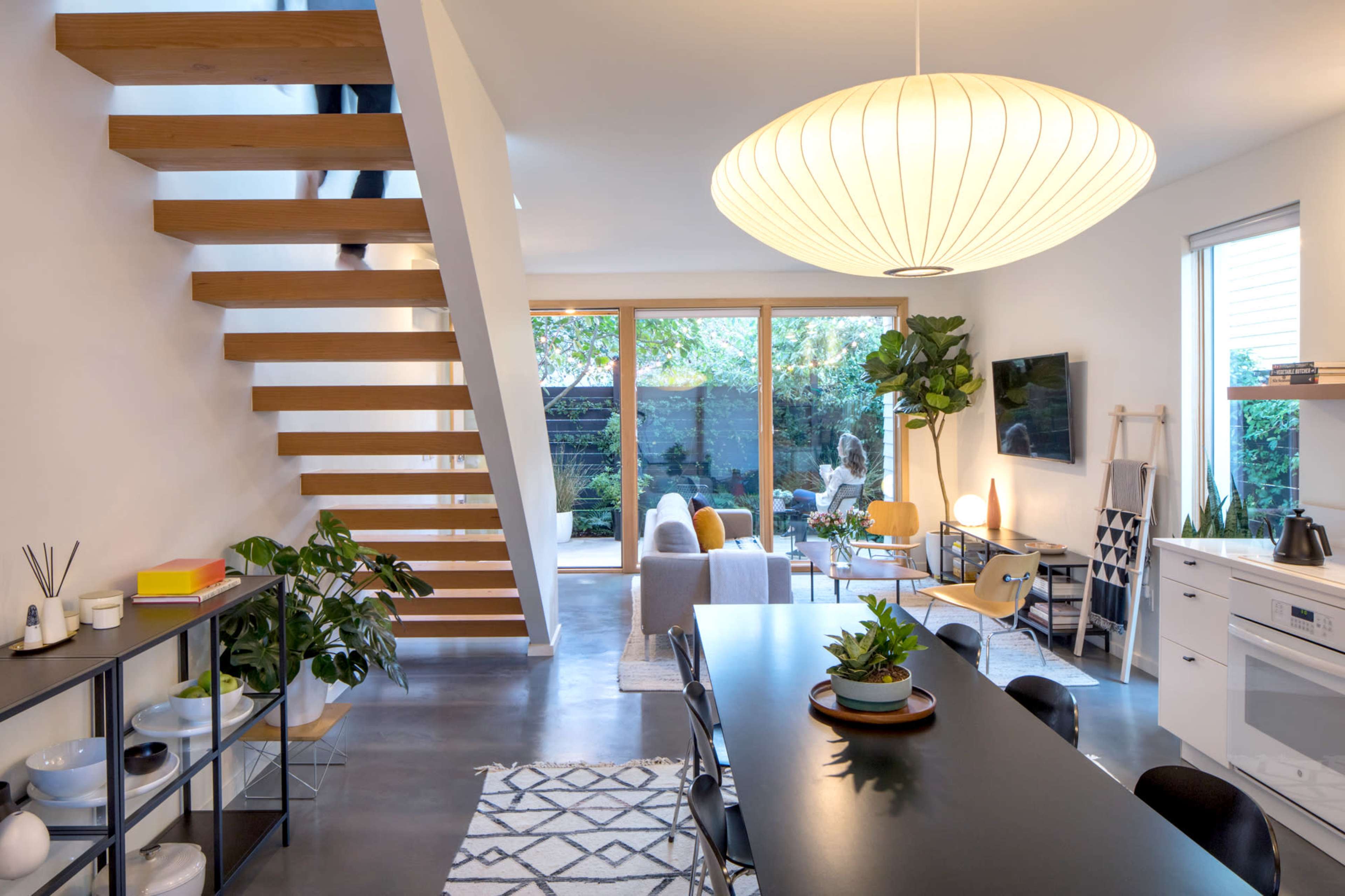 A modern dining area features a long table surrounded by black chairs, with a staircase leading to an upper level and large windows overlooking a lush outdoor space.