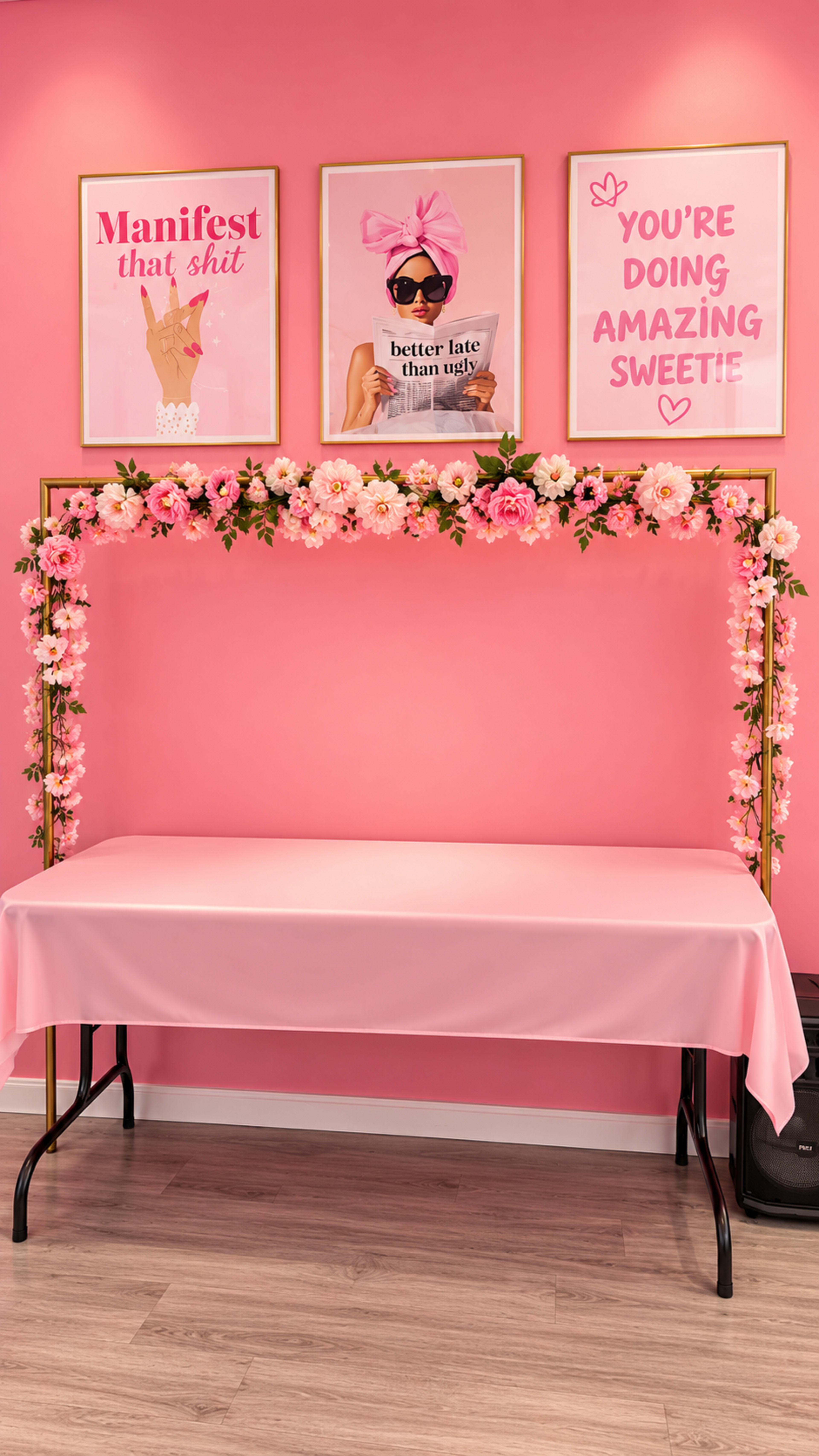 The image shows a decorative photo booth setup with a pink backdrop, a table covered in a pink tablecloth, and framed inspirational posters on the wall above.