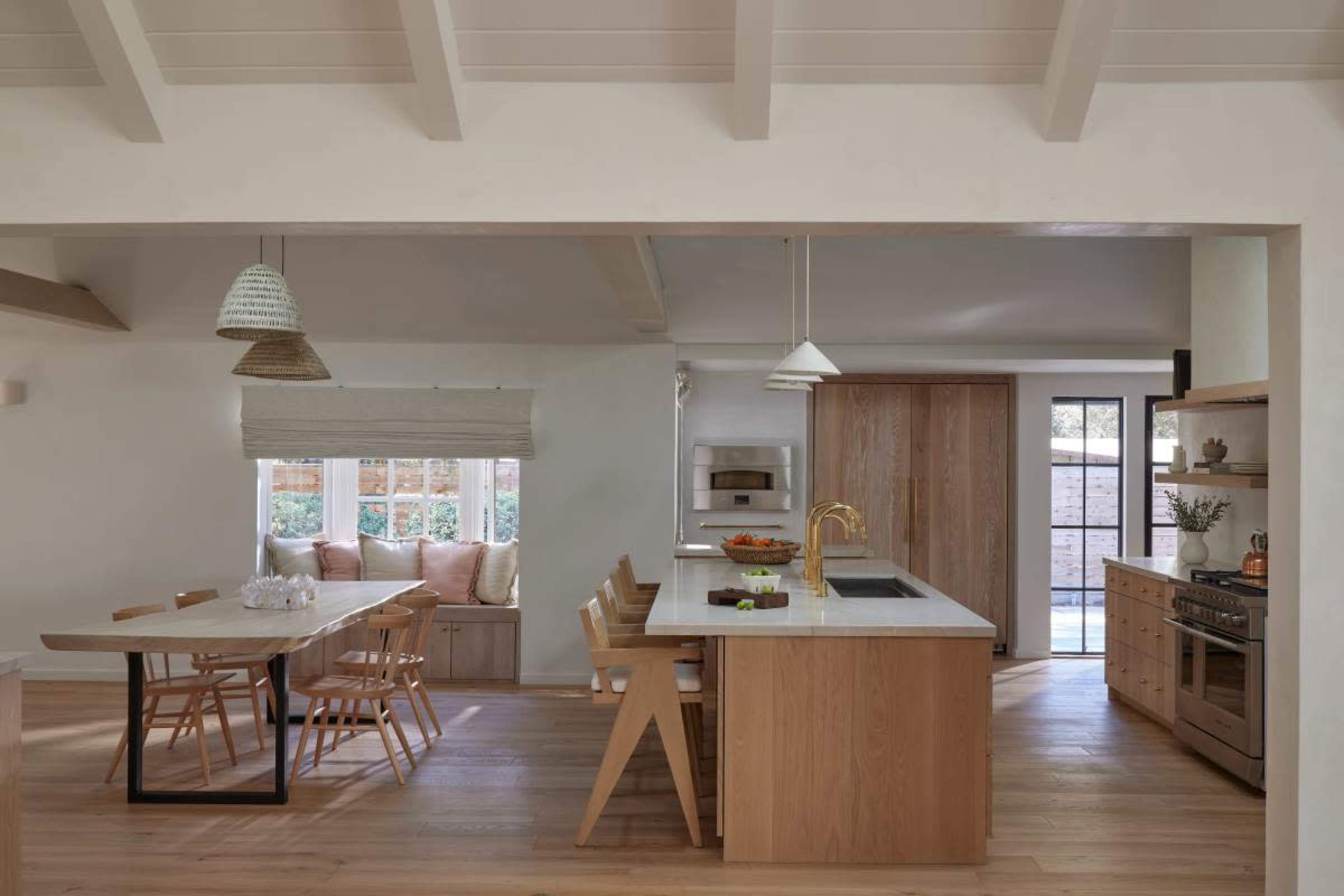 The image shows a modern kitchen with an island, wooden cabinetry, a dining table, and large windows allowing natural light to enter.