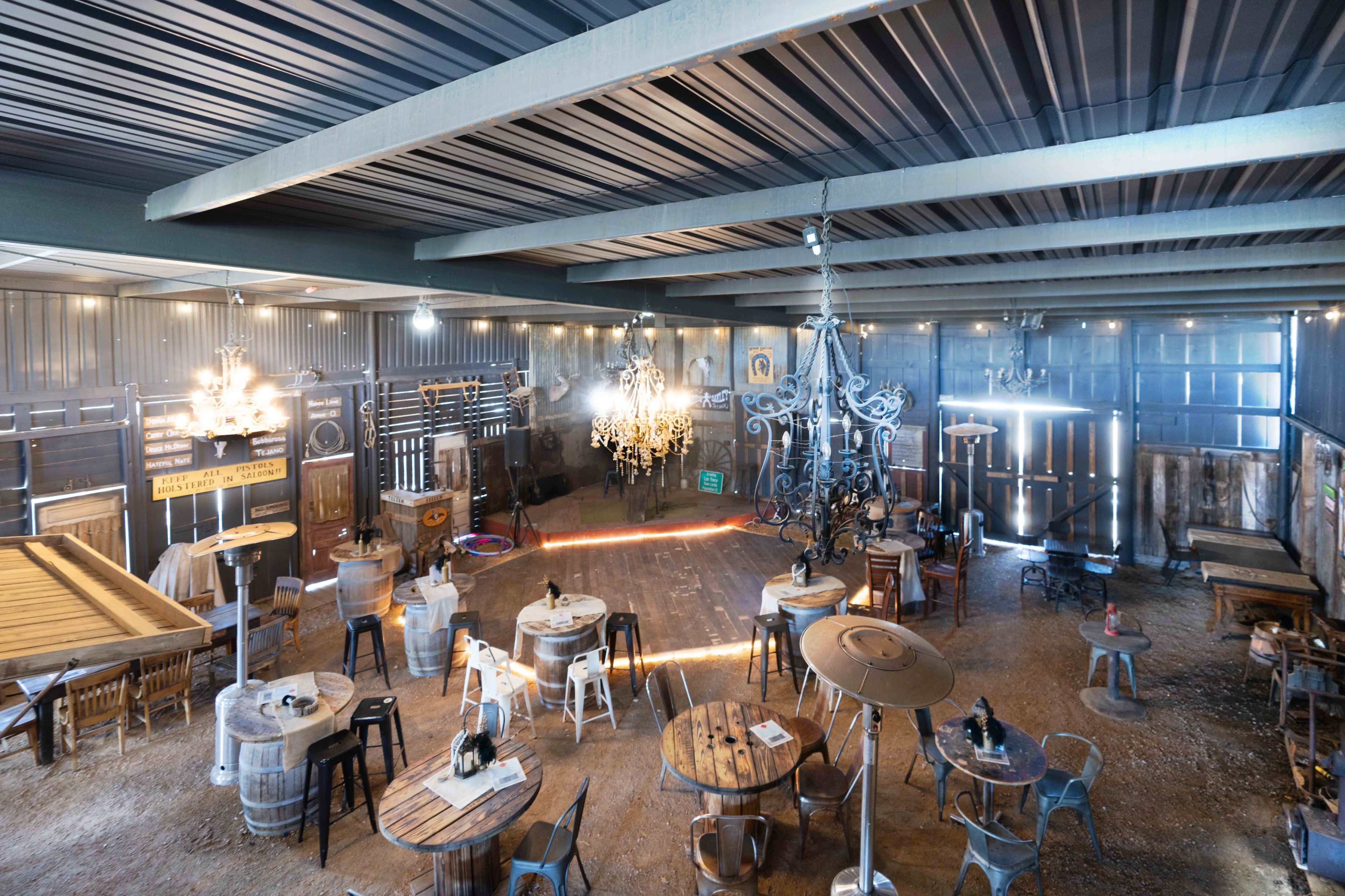 The image shows an industrial-style venue with wooden tables and chairs, chandeliers hanging from the ceiling, and a spacious dance floor illuminated by ambient lighting.