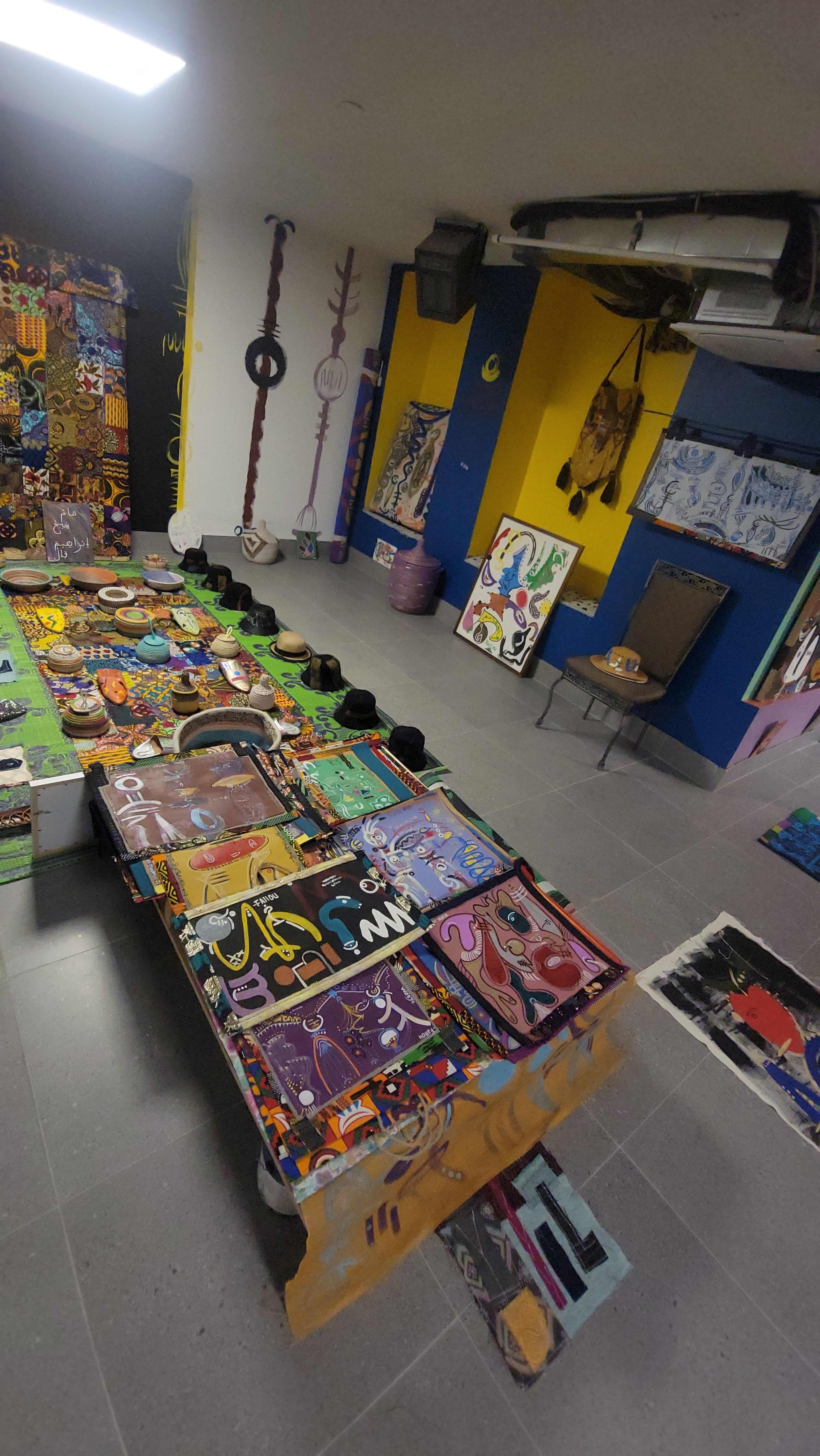 The image shows a vibrant, artistic space with a long table covered in various colorful artworks, surrounded by painted walls and additional art pieces displayed nearby.