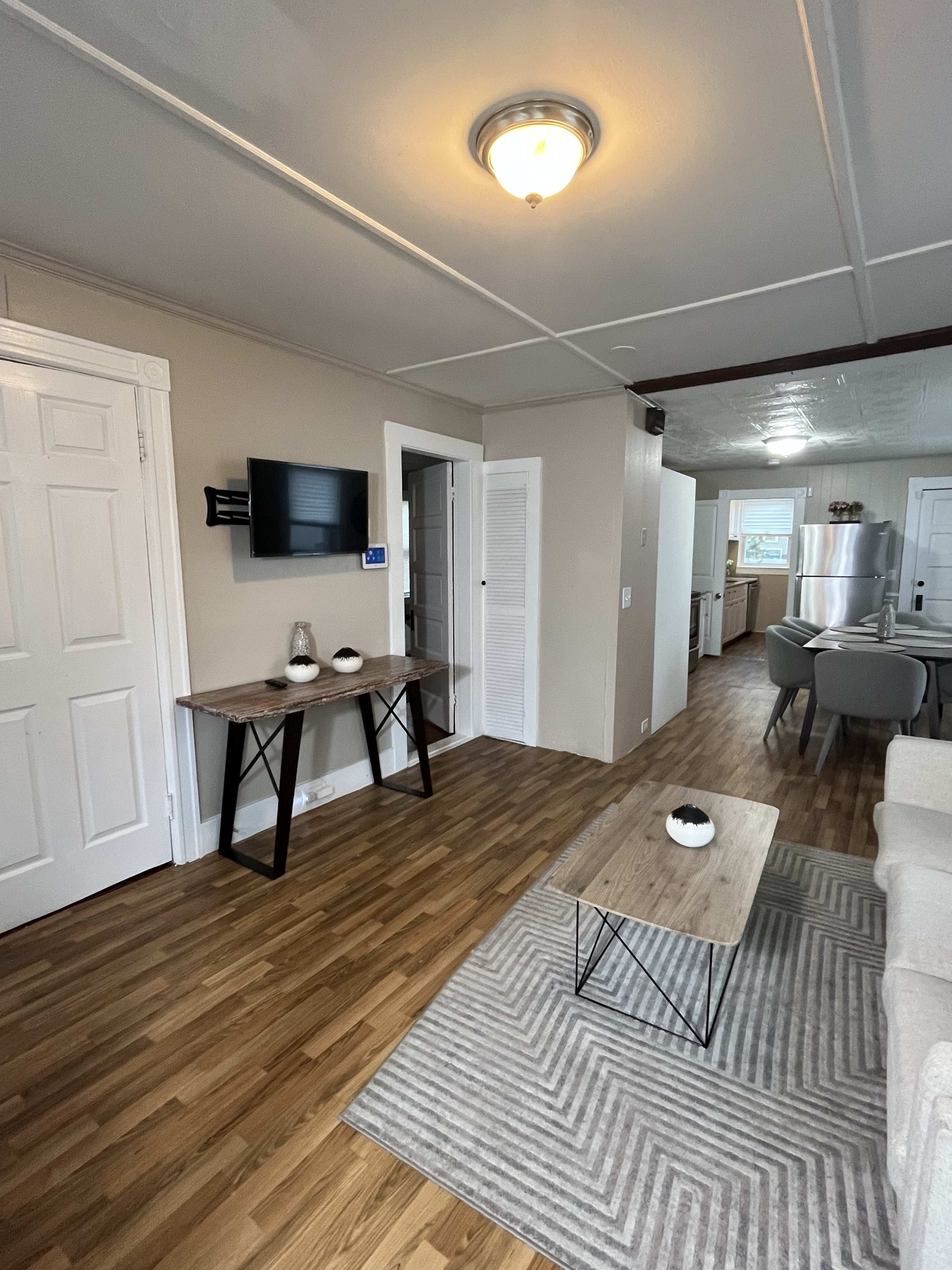 The image shows a living area featuring wooden flooring, a coffee table, a small console table with a TV mounted on the wall, and an open view into a kitchen.