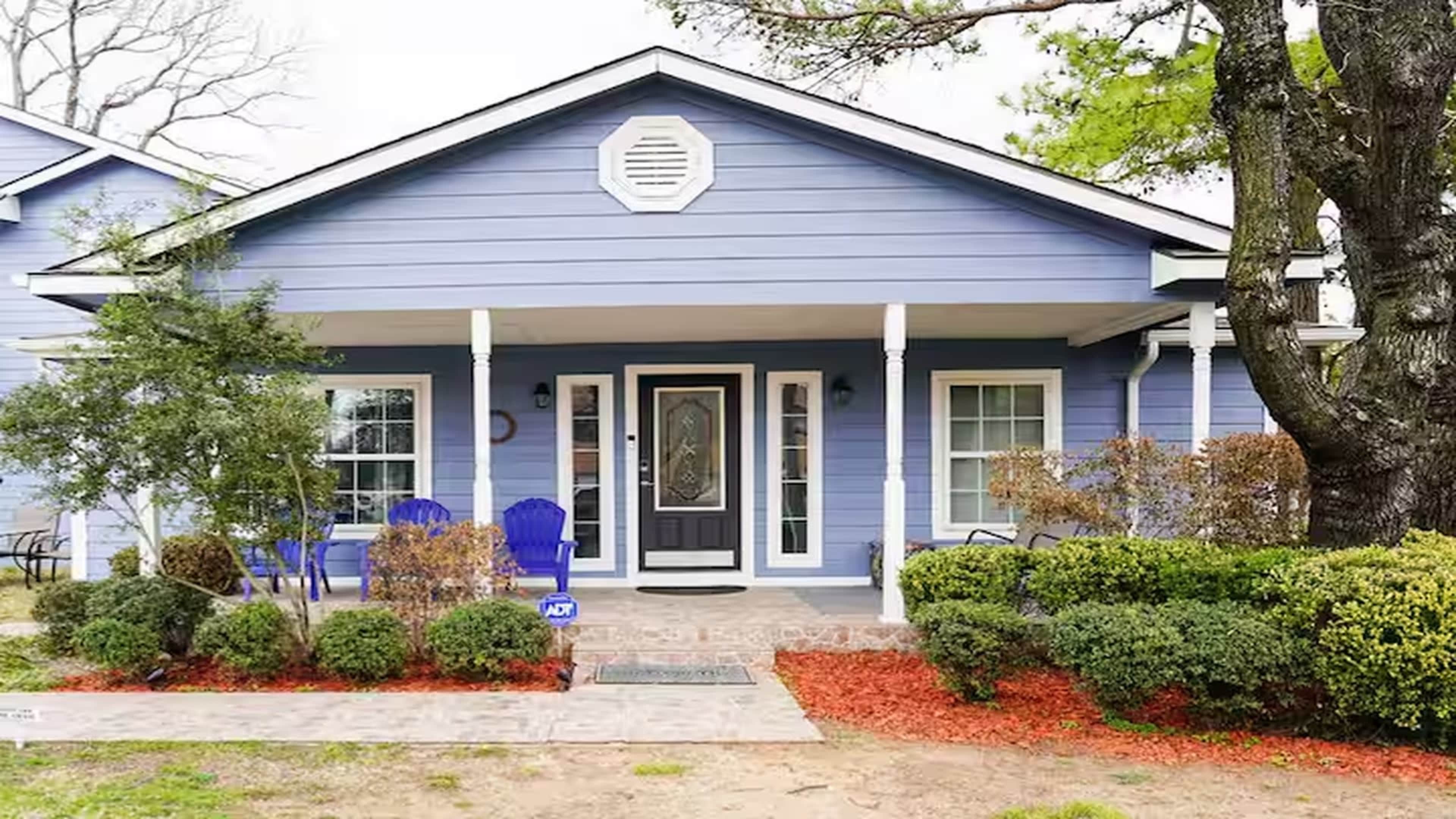 The image shows a light blue house with a neatly manicured lawn, featuring a front porch with two blue chairs and a pathway leading to the entrance.