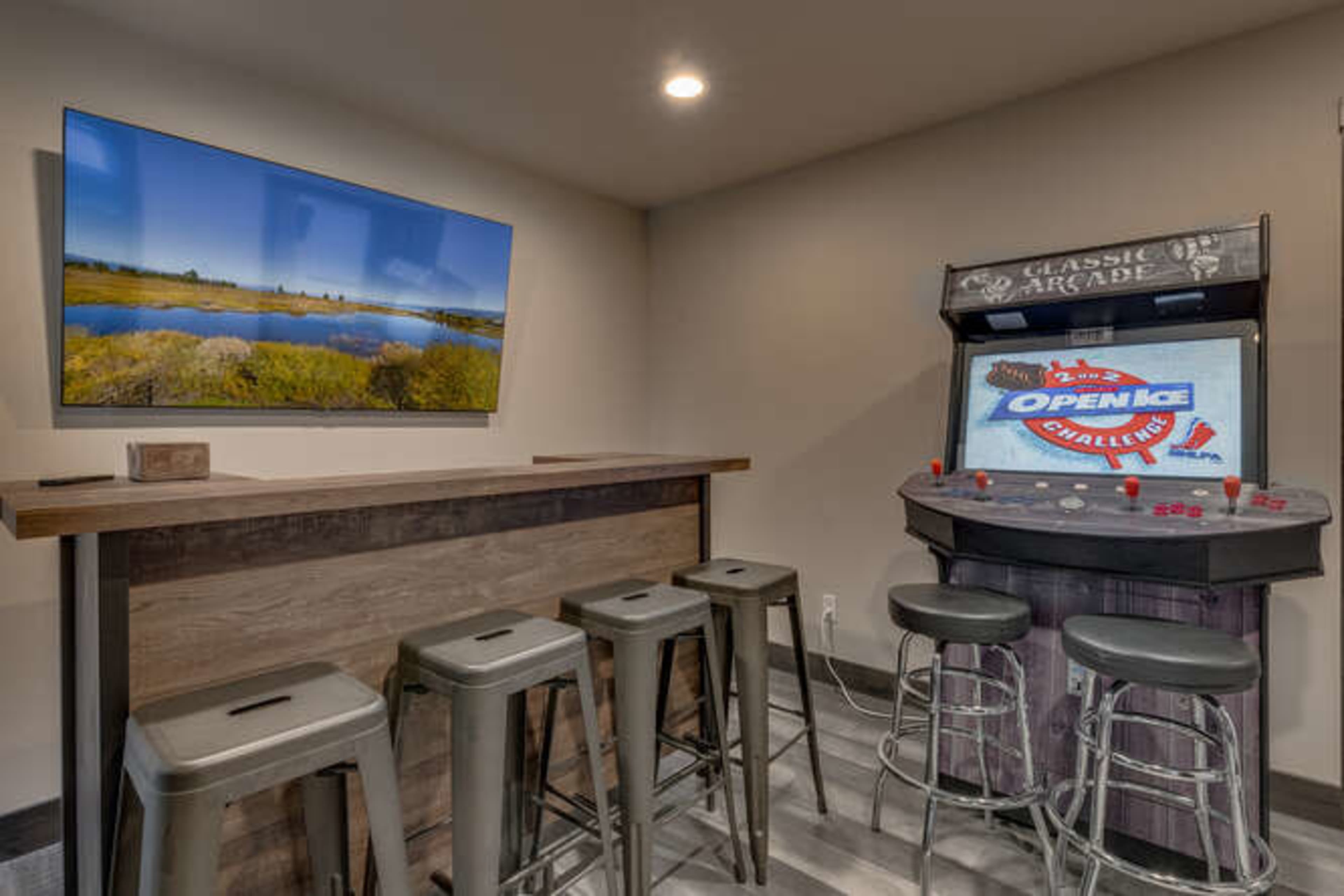 A room with a dark wooden bar and four metal stools, featuring an arcade game machine against the wall with a large television displaying a landscape photograph.