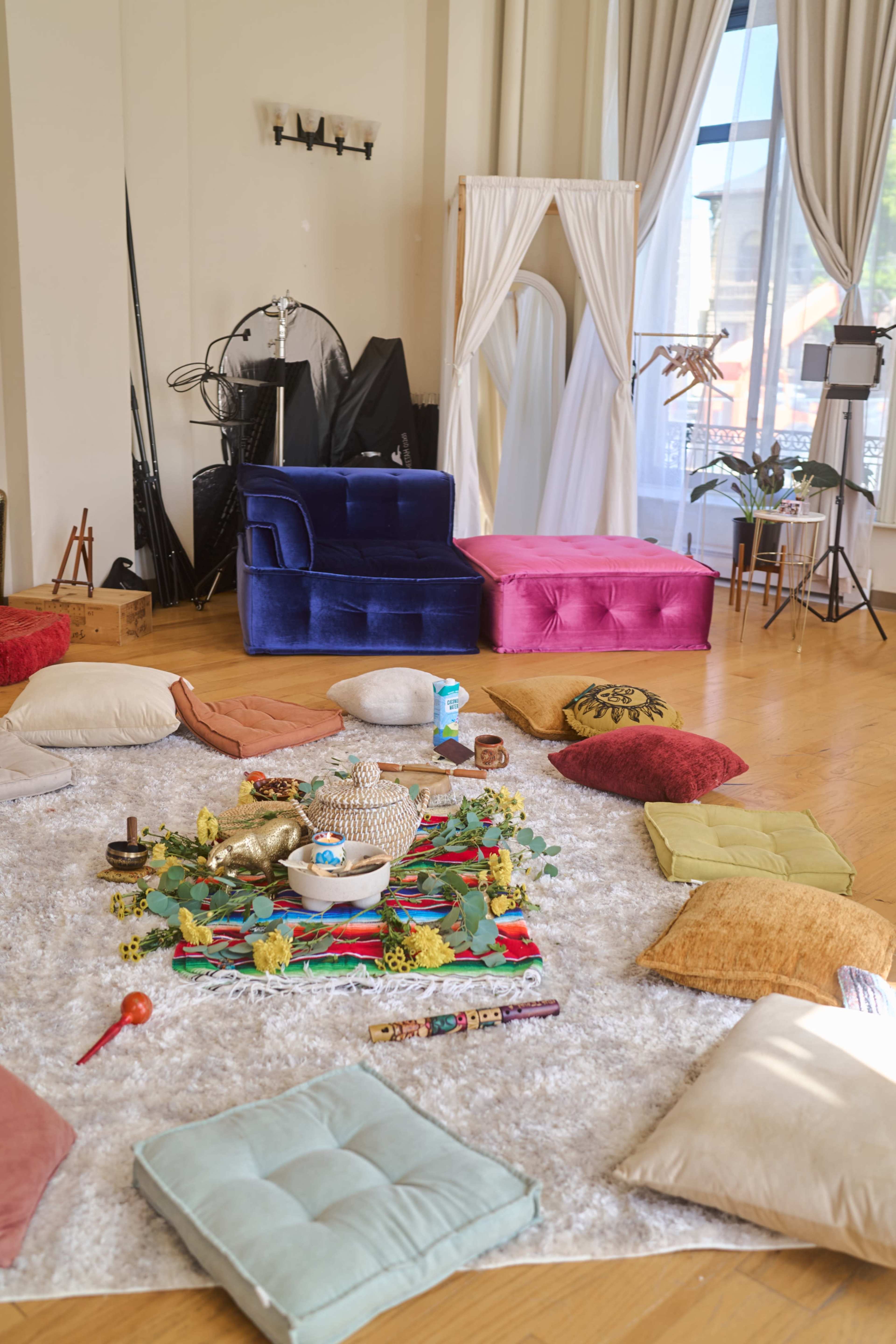 The image shows a spacious room with various seating cushions arranged on a soft rug, surrounded by colorful furniture and decorative items.