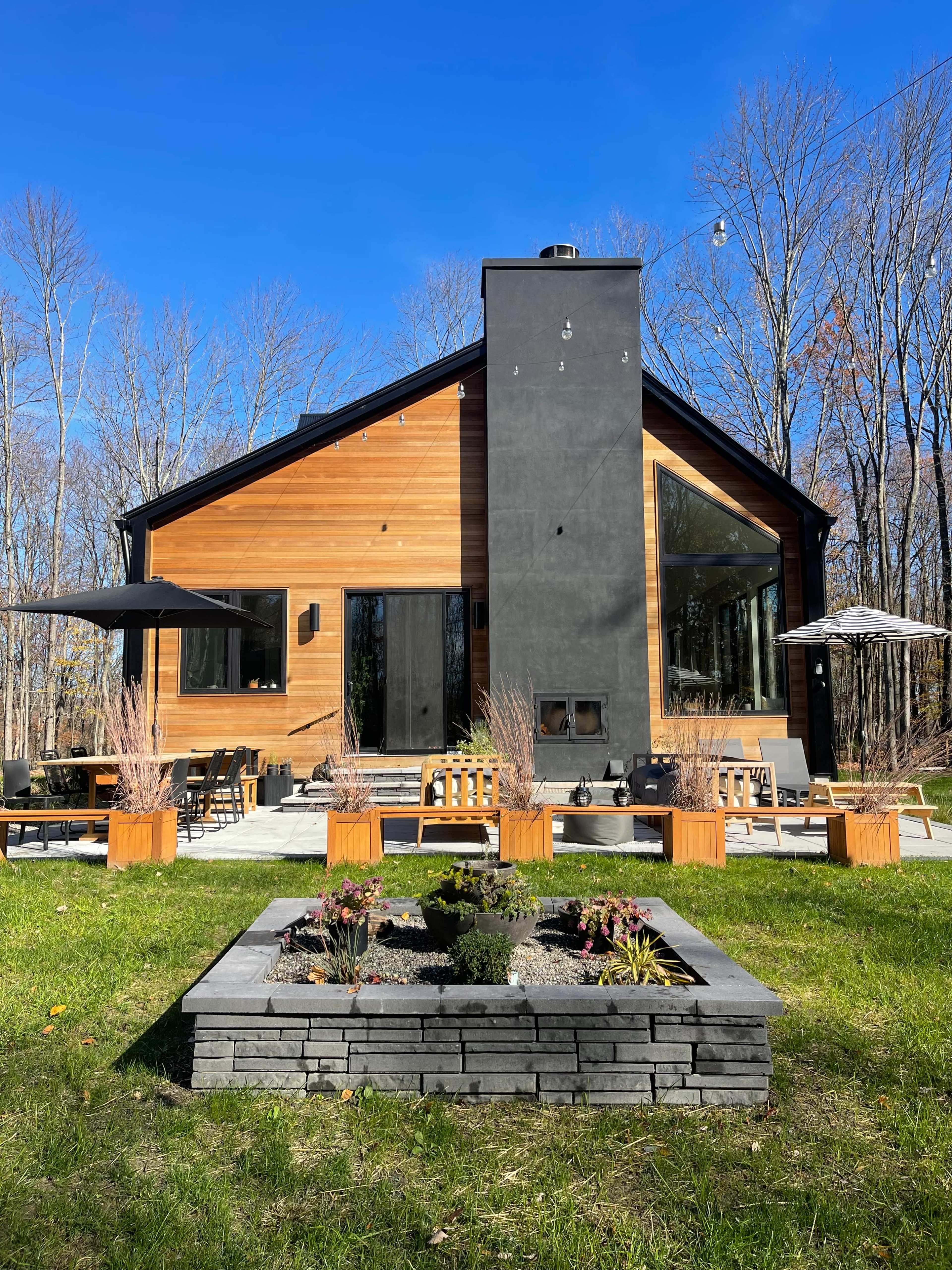 A modern house with a wooden exterior and a prominent black chimney, set against a backdrop of trees and a landscaped garden featuring a stone planter.