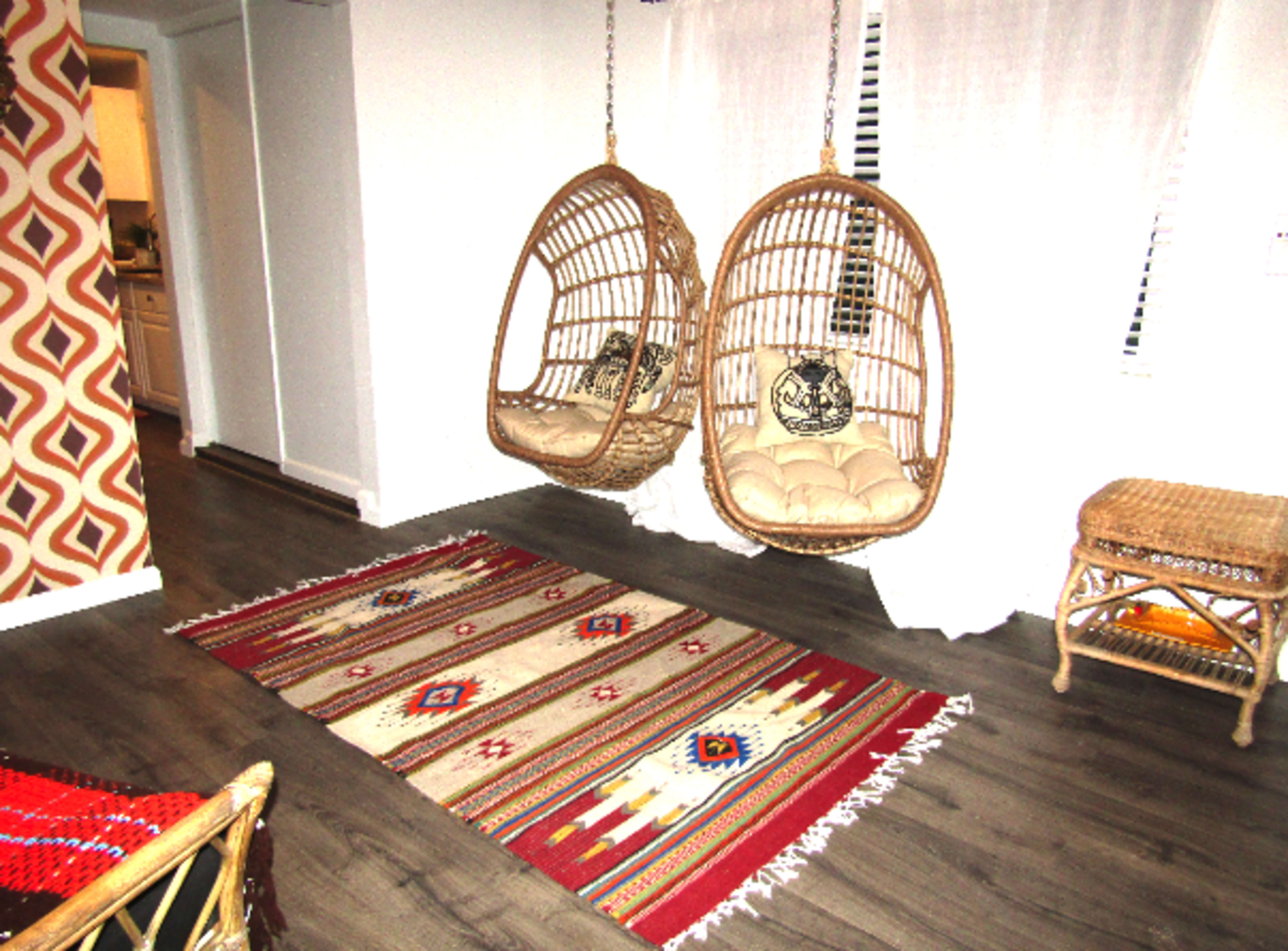 The image shows a living space featuring two hanging wicker chairs beside a patterned rug on a wooden floor.
