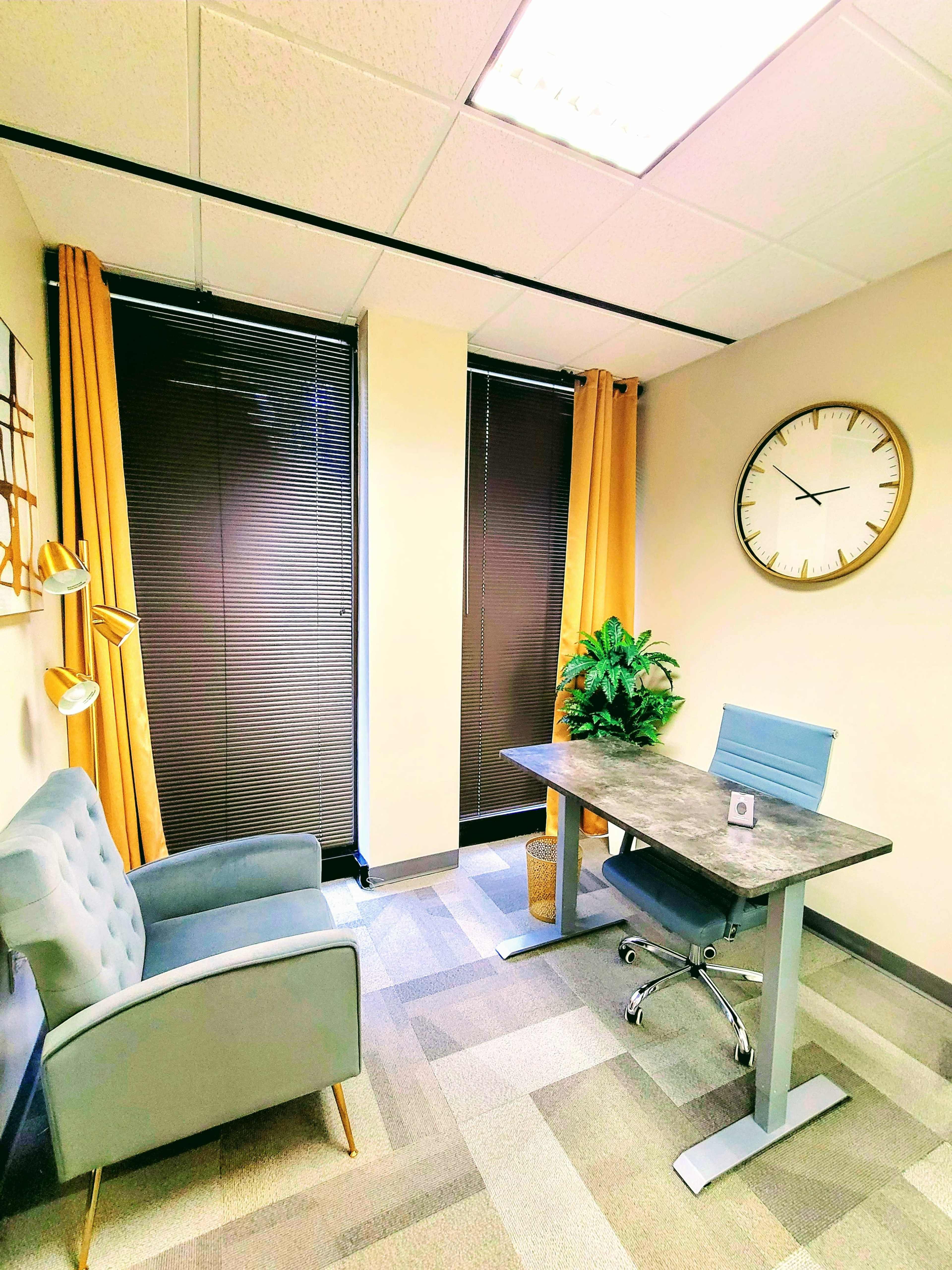A modern office space featuring a gray desk, a blue chair, a small plant, and a comfortable armchair, with large windows covered by blinds and yellow curtains.
