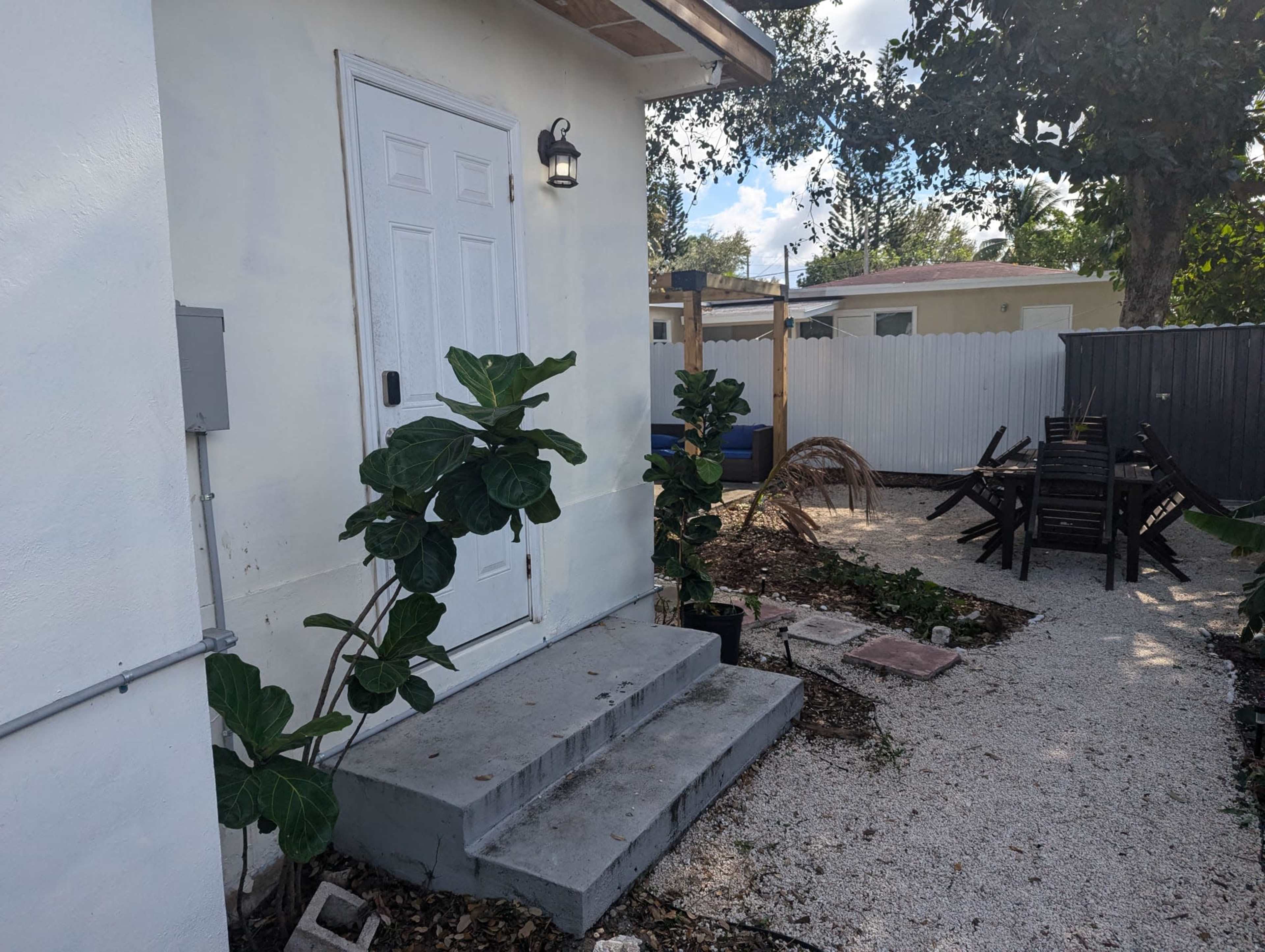 The image shows a small white building with a concrete entryway, surrounded by green plants and a gravel pathway leading to a backyard area with outdoor furniture.