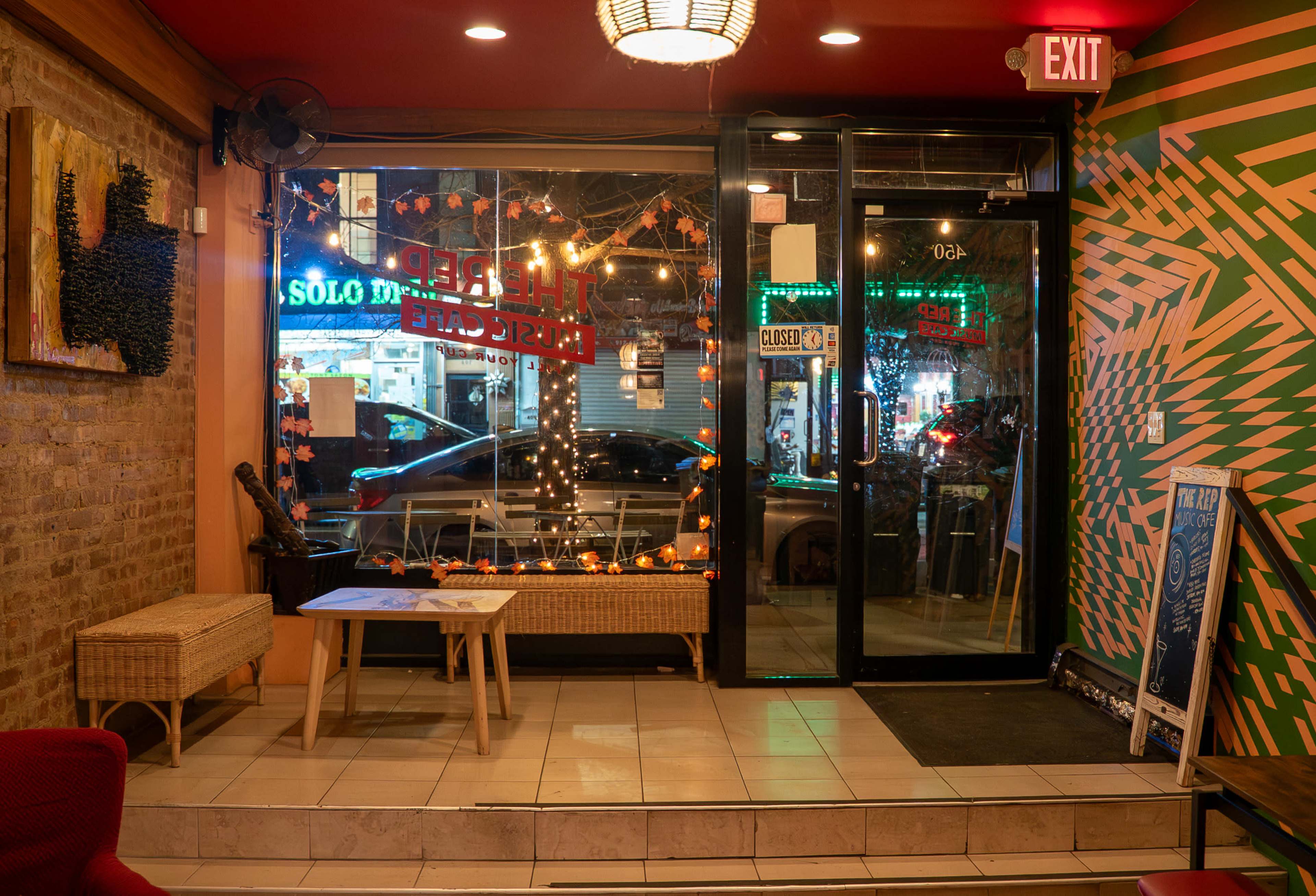 The image shows the interior of a café with a view of a brightly lit street through large windows, featuring decorative lights and a cozy seating area.