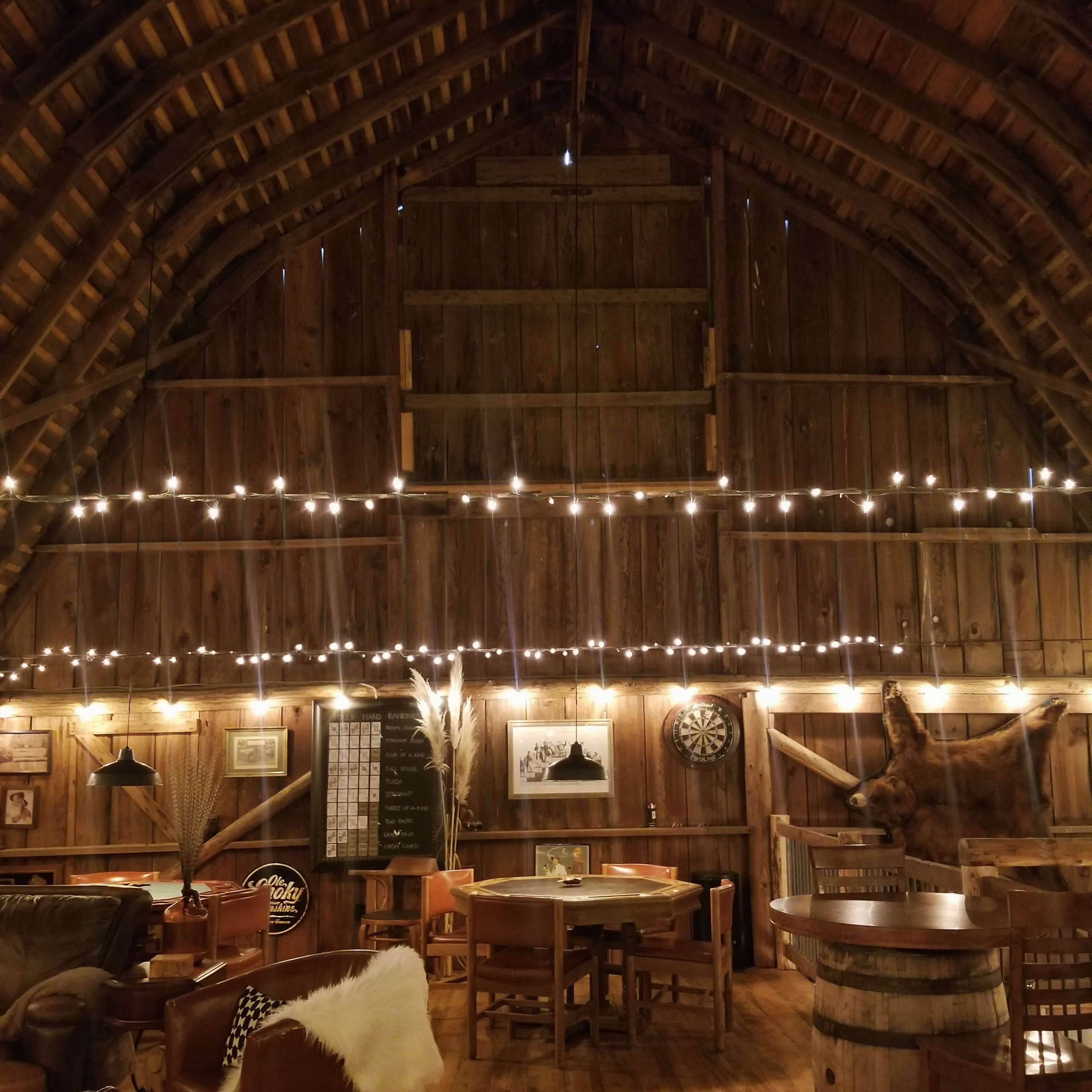 The interior of a rustic barn features wooden beams, string lights, and various seating arrangements.