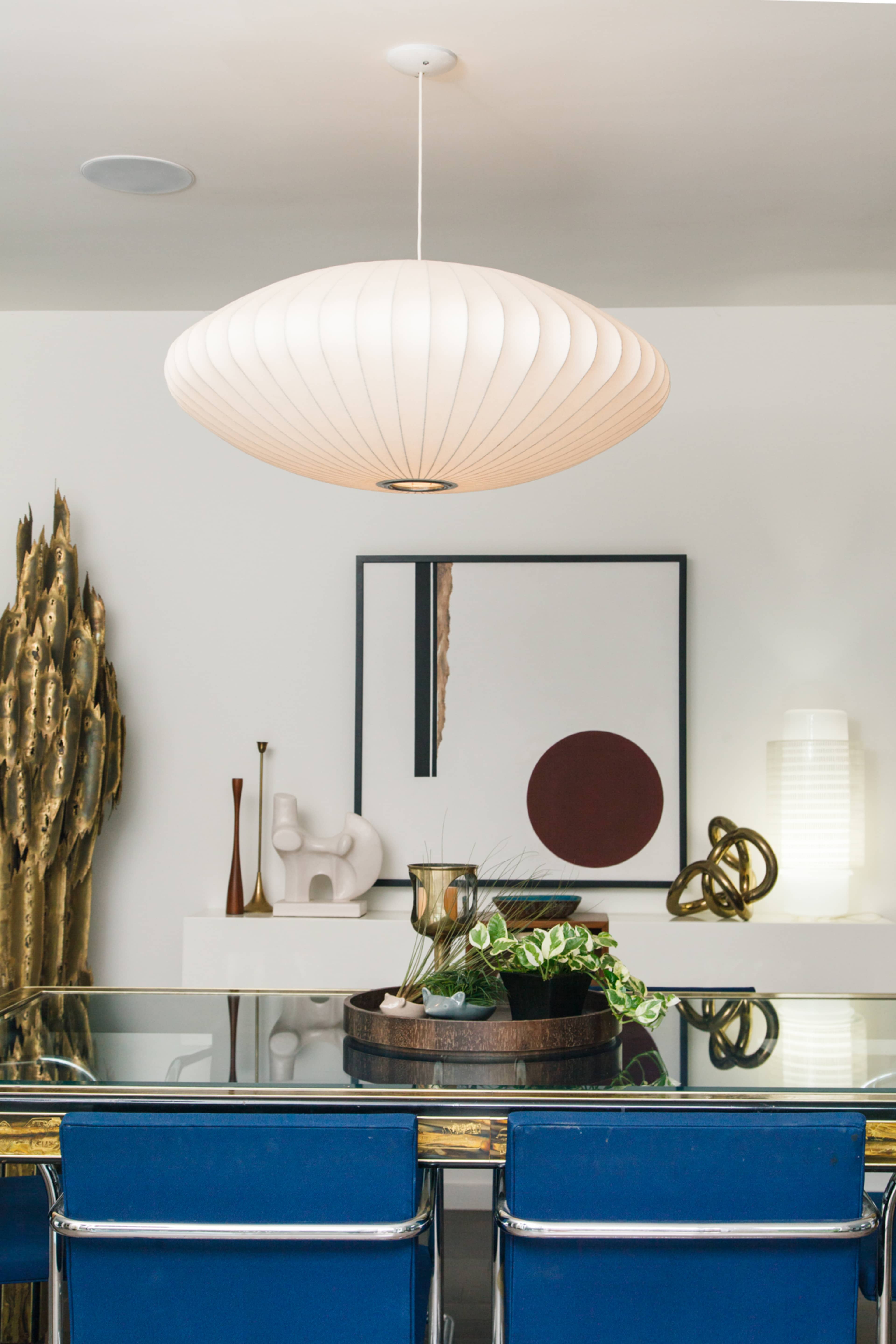 A modern dining area features a round pendant light, a glass table with blue chairs, and decorative elements including a plant arrangement and abstract art on the wall.
