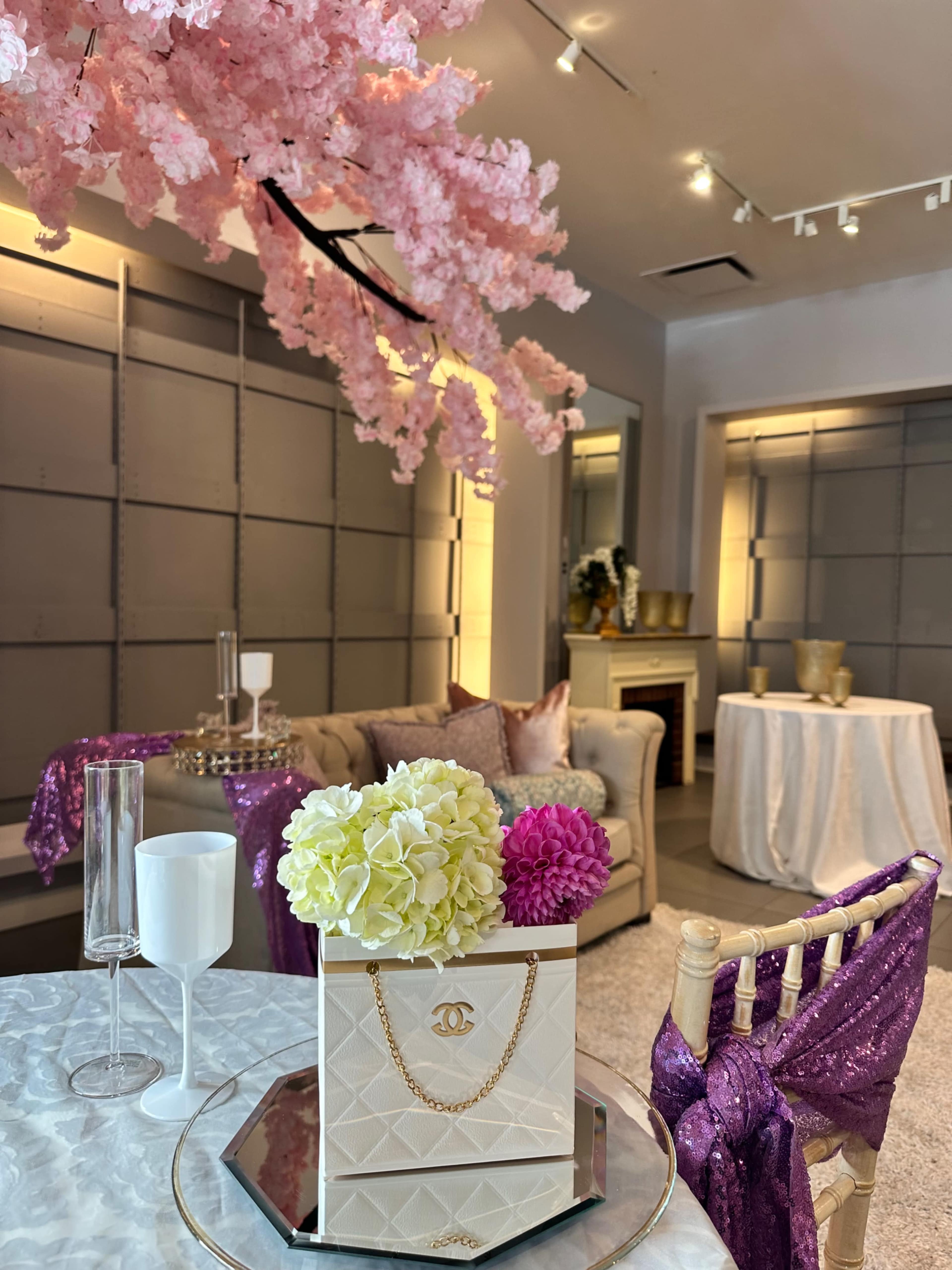 The scene features a stylish interior with a decorative table, a Chanel bag filled with flowers, and cherry blossom branches overhead.
