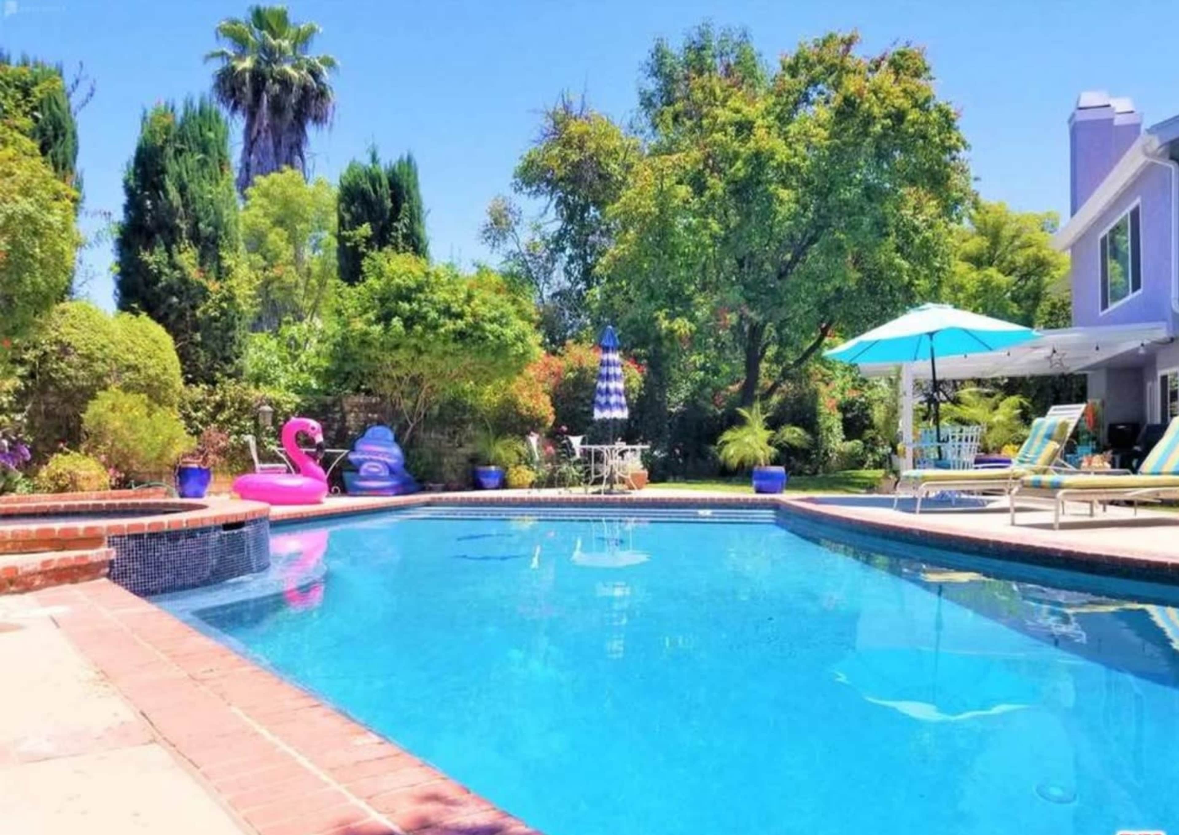 A clear swimming pool is surrounded by green trees, patio furniture, and a pink flamingo float.