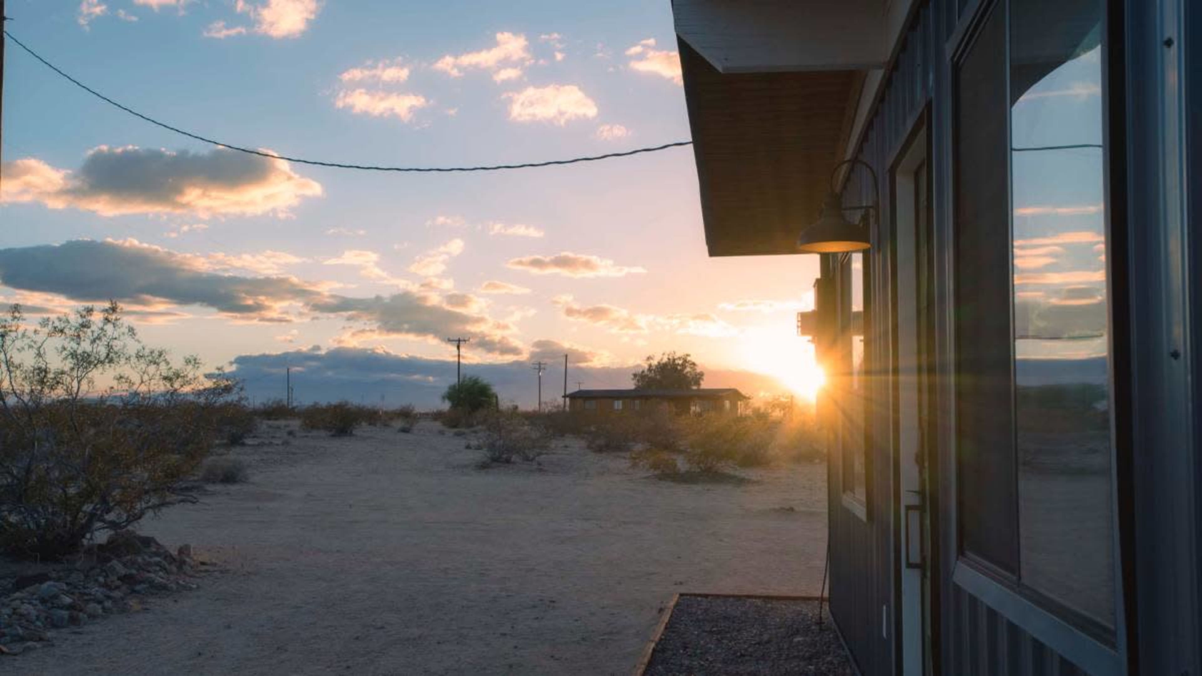 The image shows a desert landscape at sunset, featuring a structure on the right side with a window reflecting the sunlight.
