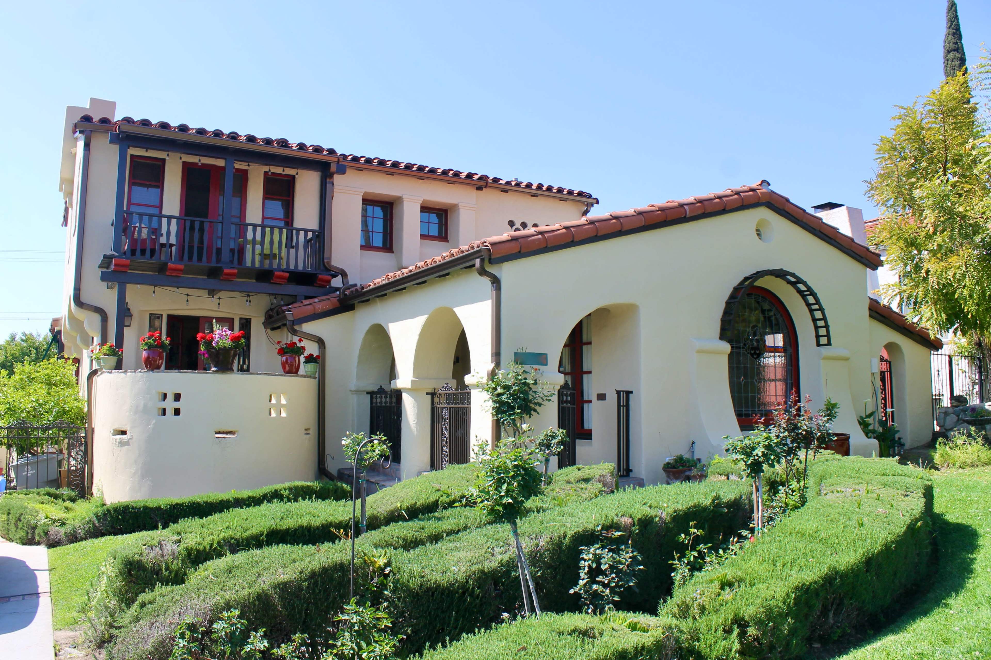 A two-story house with a red-tiled roof, arched windows, and manicured shrubs in the front yard.