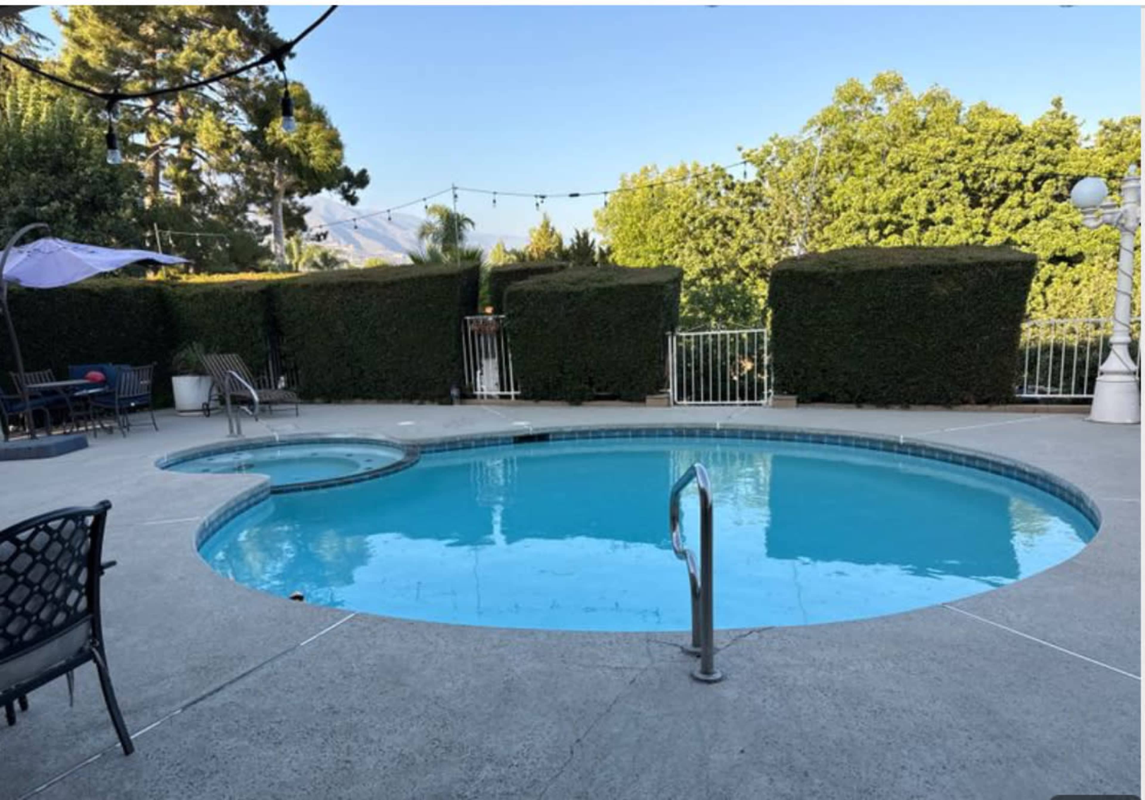 A clear, circular swimming pool with a connected hot tub is surrounded by neatly trimmed hedges and outdoor seating.