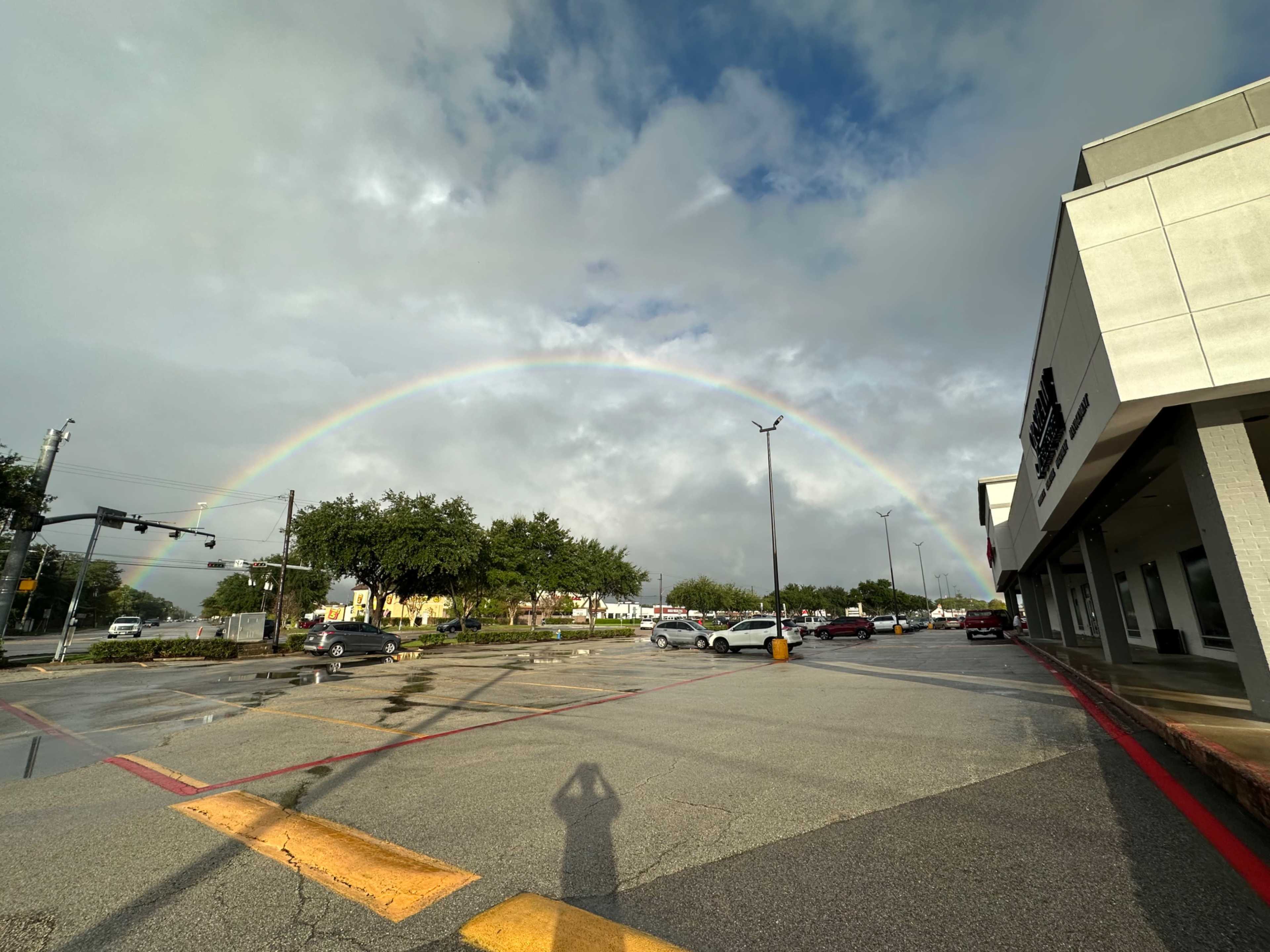 A rainbow arches over a parking lot after rain, with partially cloudy skies and vehicles parked nearby.