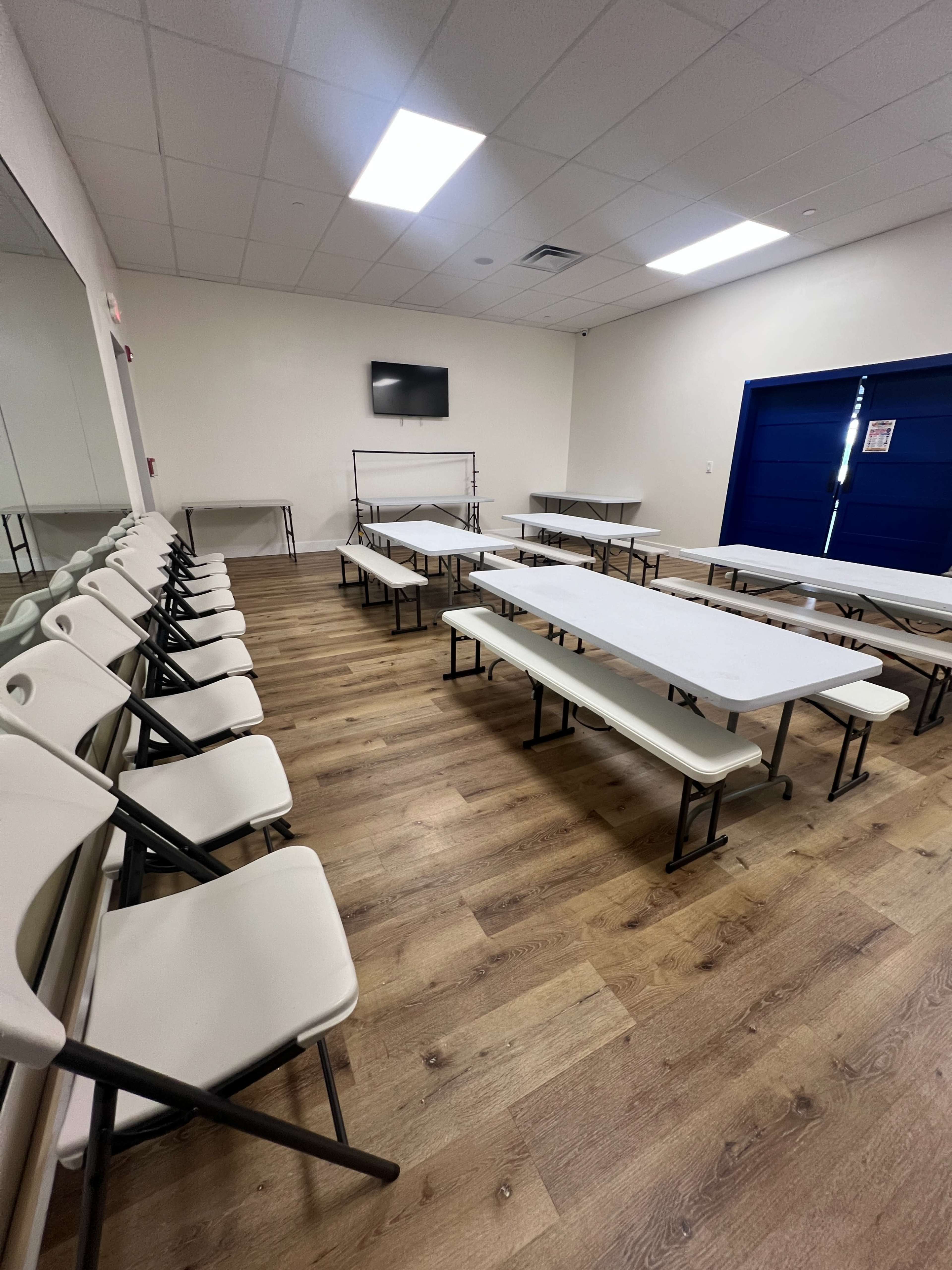 The image shows a large, empty room with multiple folding tables arranged in rows and chairs positioned around them, along with a television mounted on the wall.