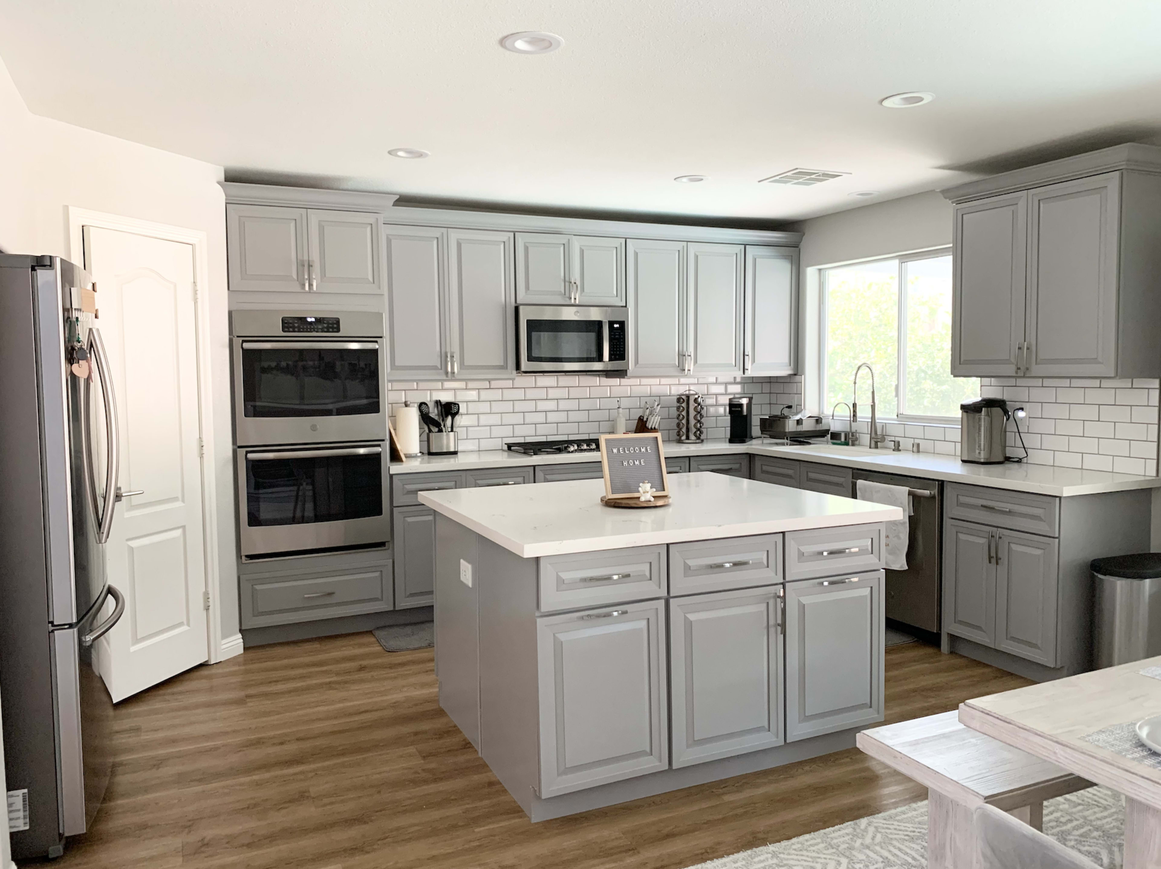 The kitchen features gray cabinets, stainless steel appliances, a central island with seating, and a window providing natural light.