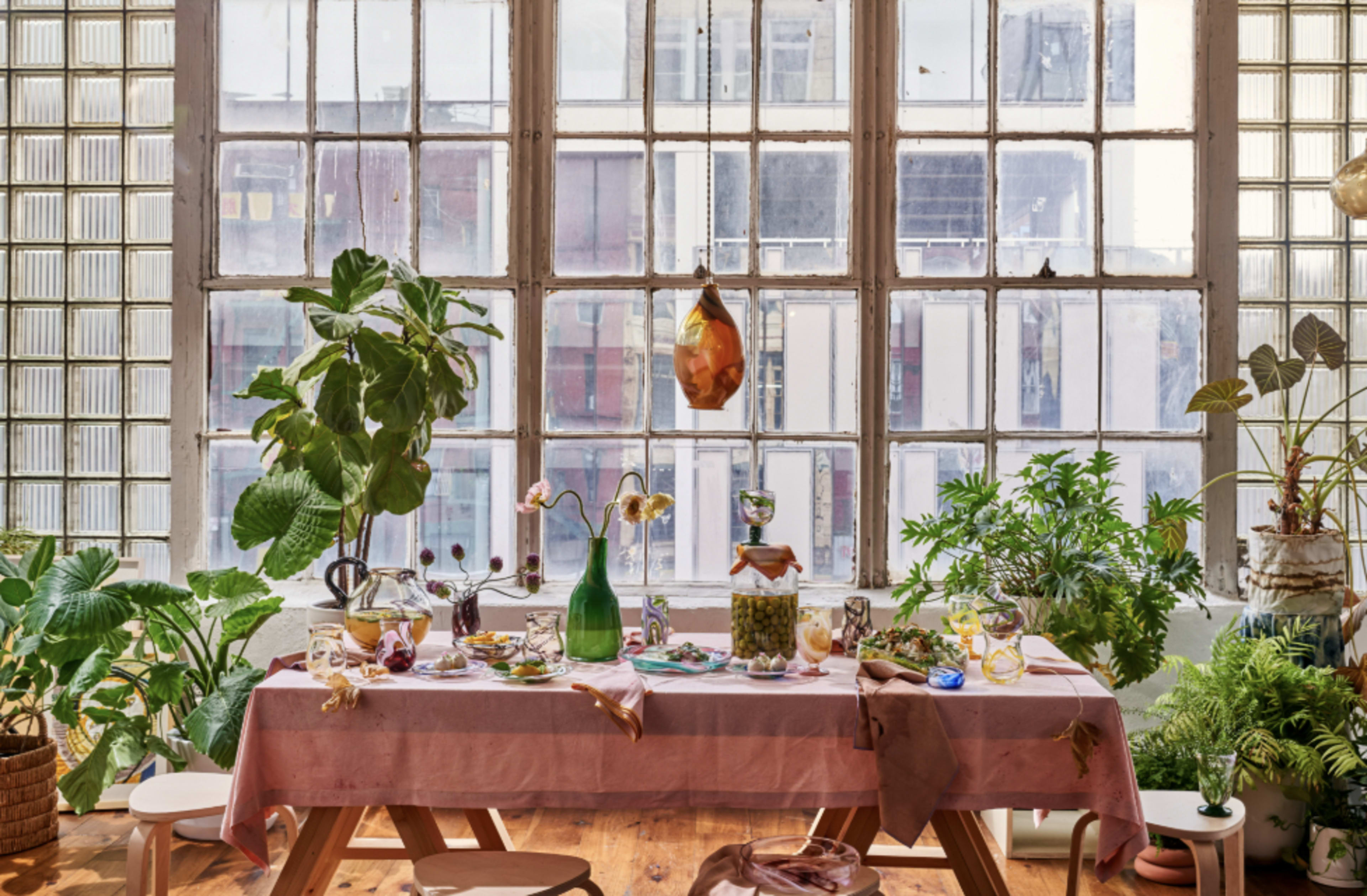 A dining table adorned with various beverages and plants is set against a backdrop of large windows with natural light.