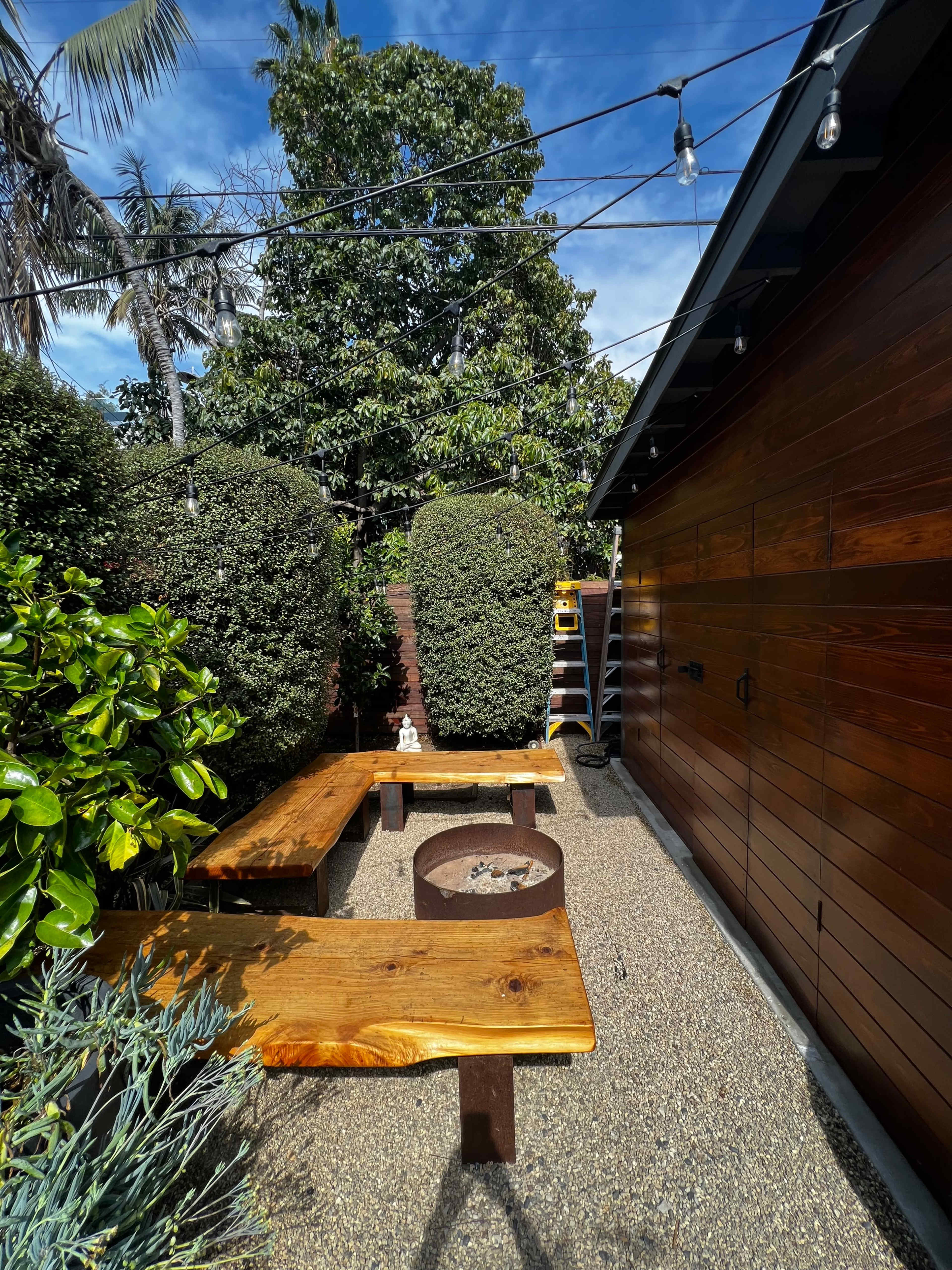 The scene features a backyard area with two wooden benches surrounding a circular fire pit, flanked by lush greenery and a wooden shed.