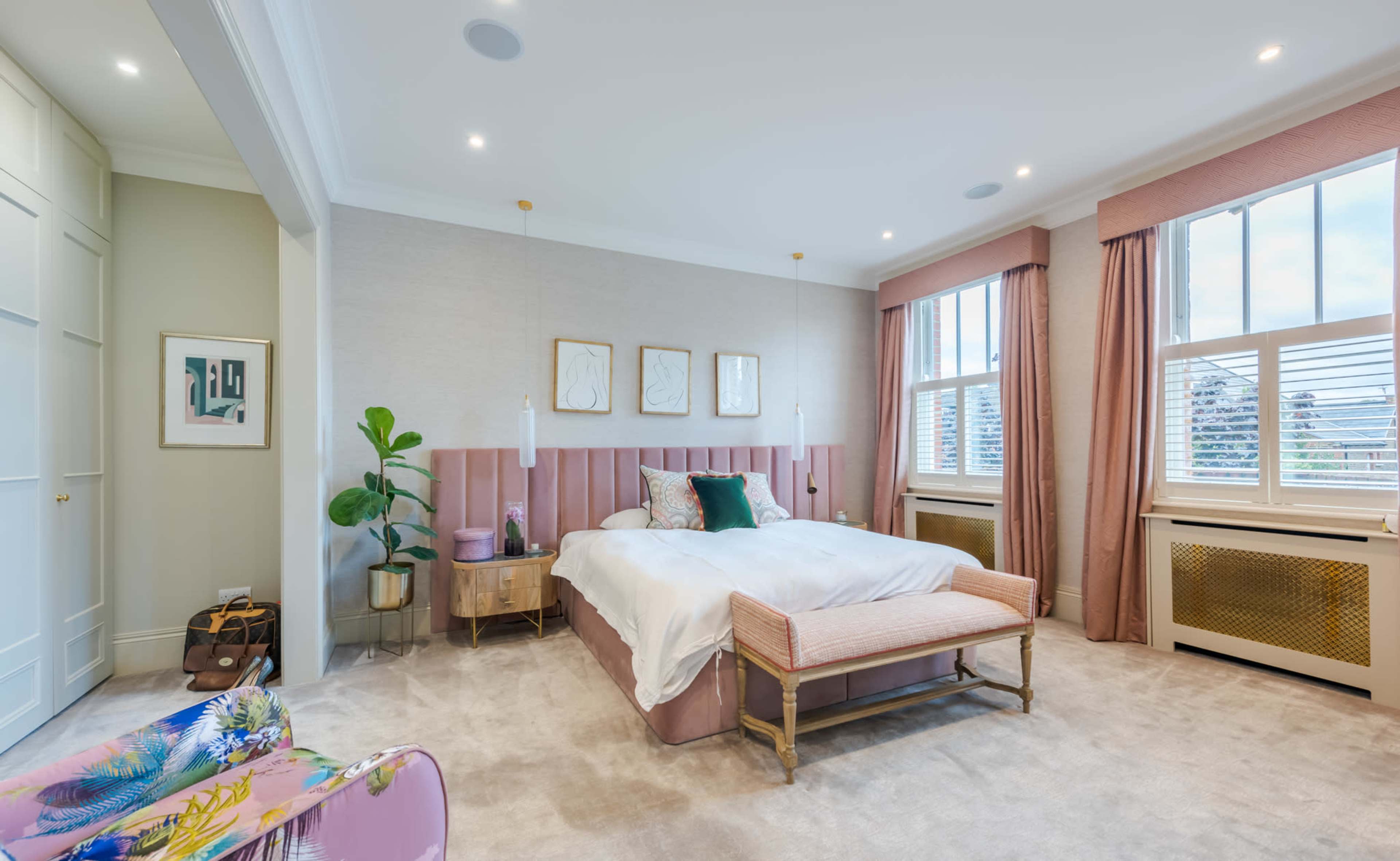 The image shows a well-decorated bedroom featuring a pink upholstered headboard, a light-colored bedspread, and a bench at the foot of the bed, with large windows allowing natural light to enter.