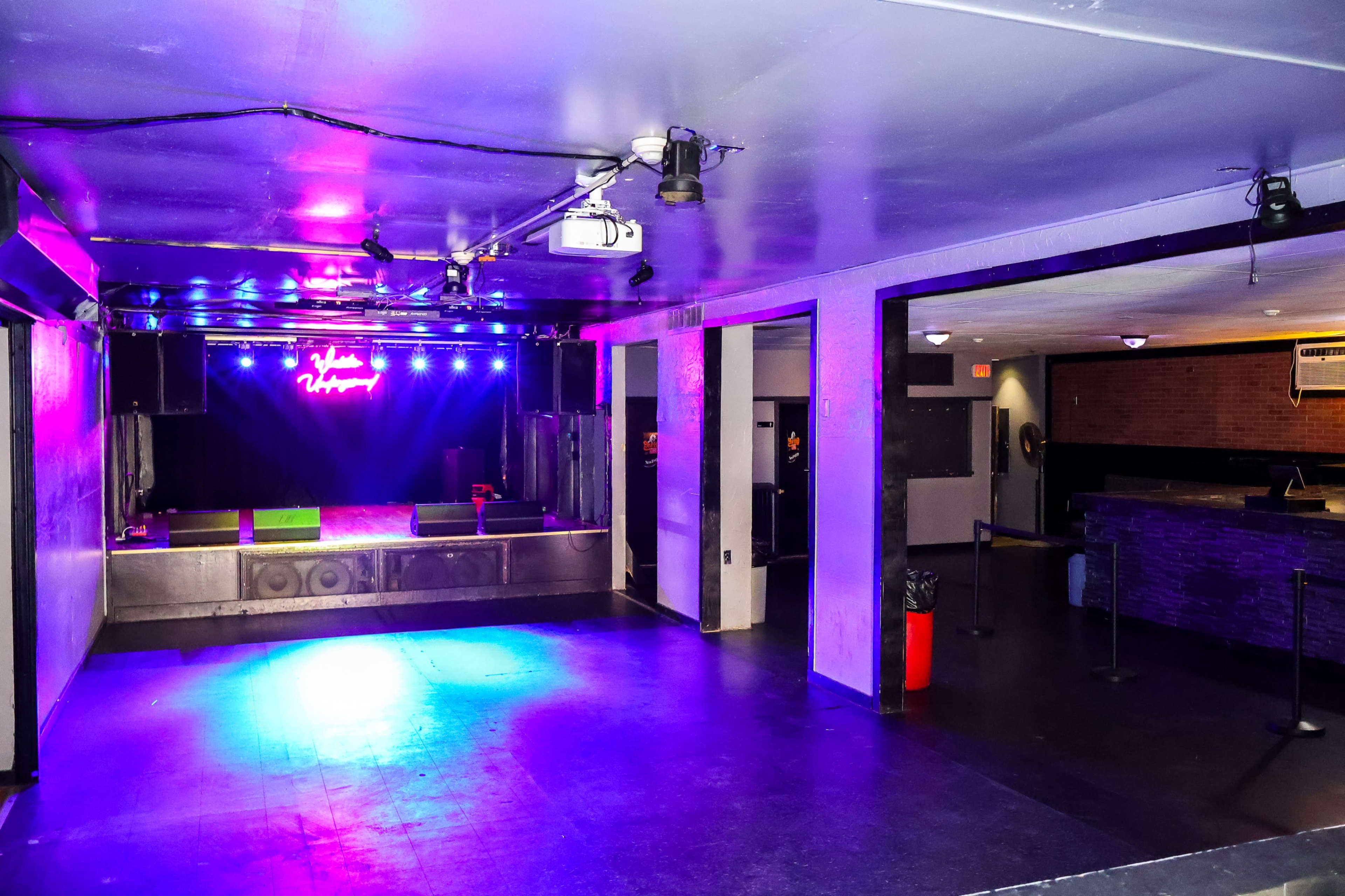 An empty music venue with a stage illuminated by colorful lights and a bar area in the background.