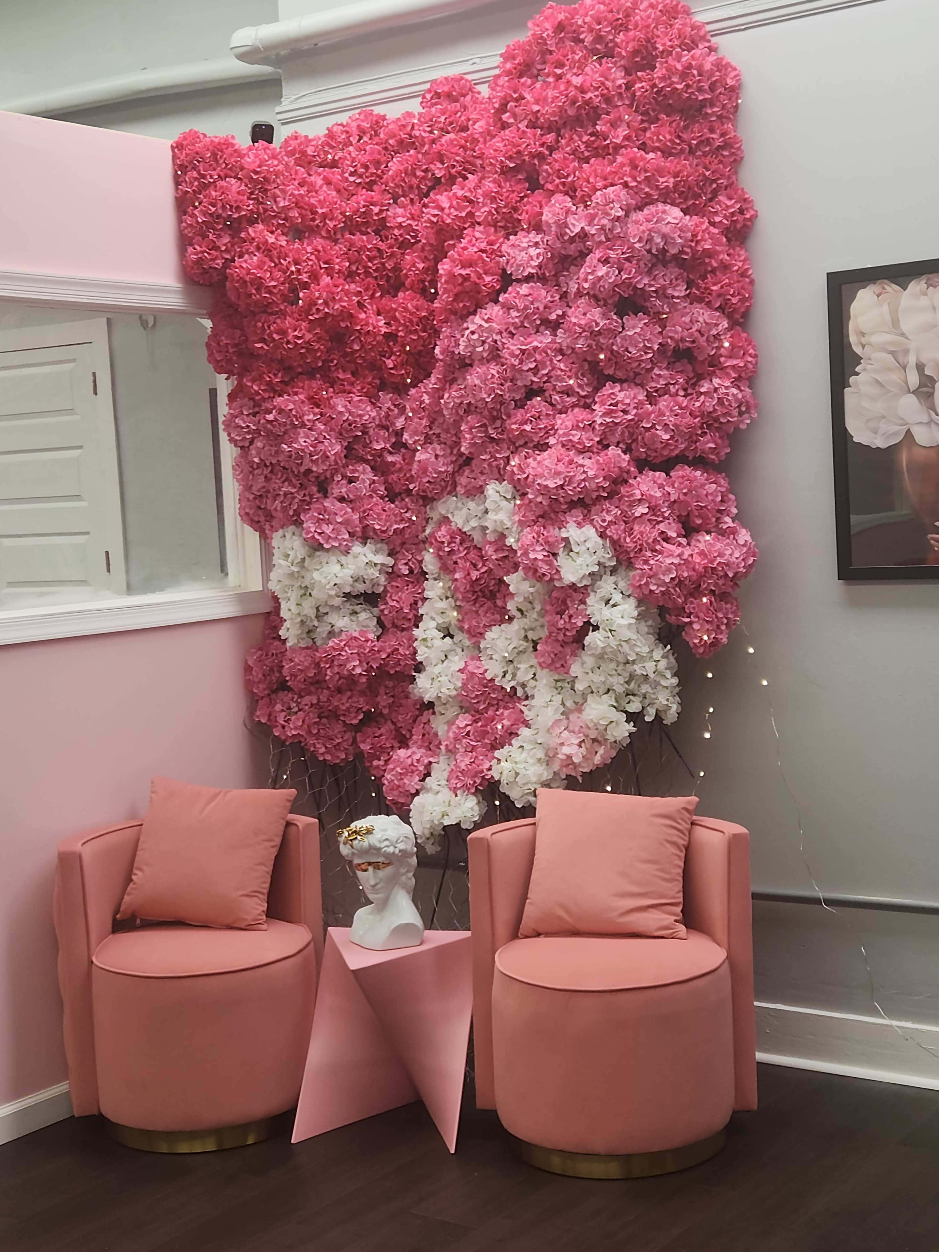 The scene features a stylish seating area with two pink upholstered chairs and a geometric table, adorned by a large wall of artificial pink and white flowers.