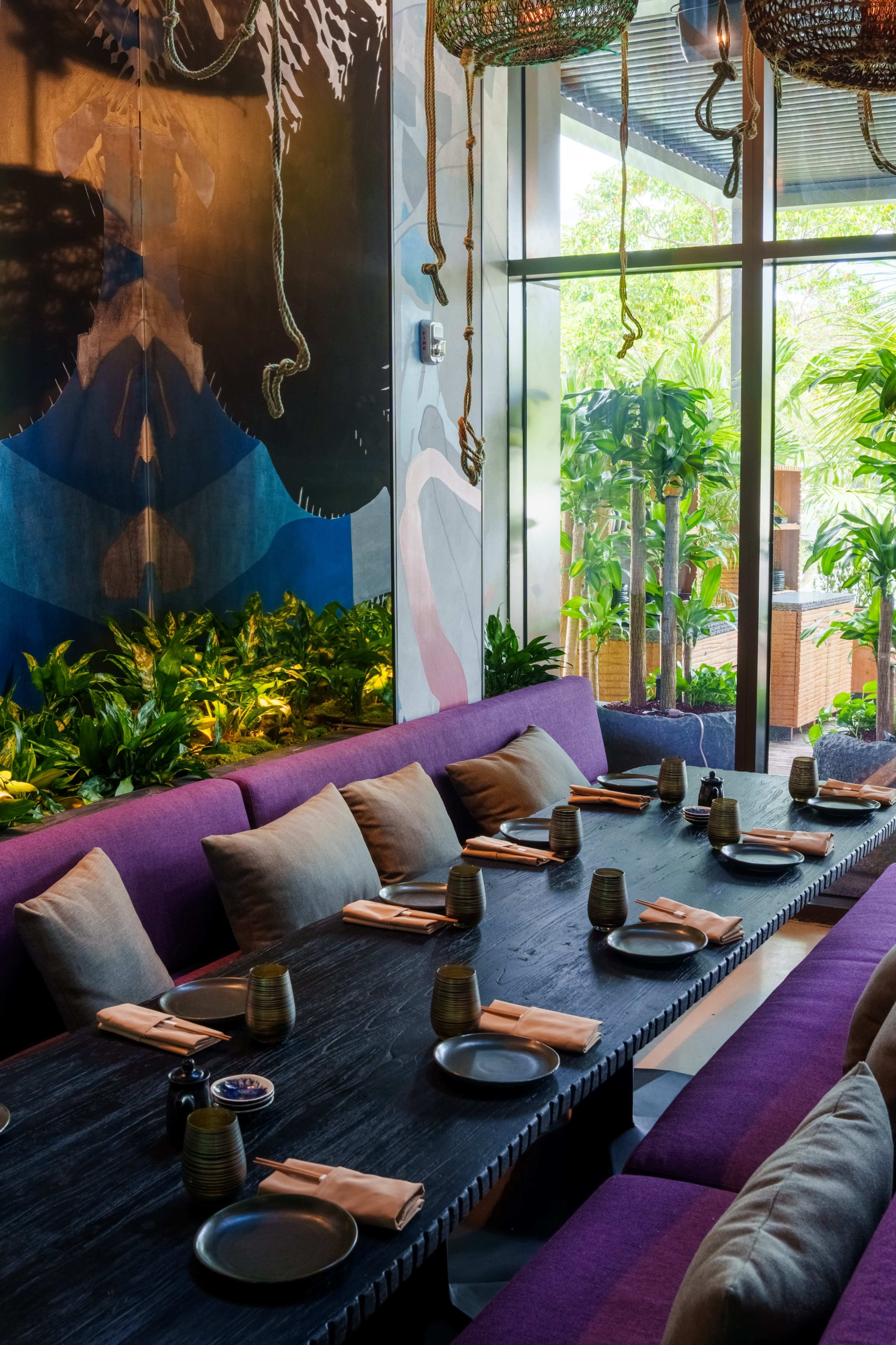 A modern restaurant interior features a long table set with napkins and tableware, surrounded by lush greenery and artistic wall murals.