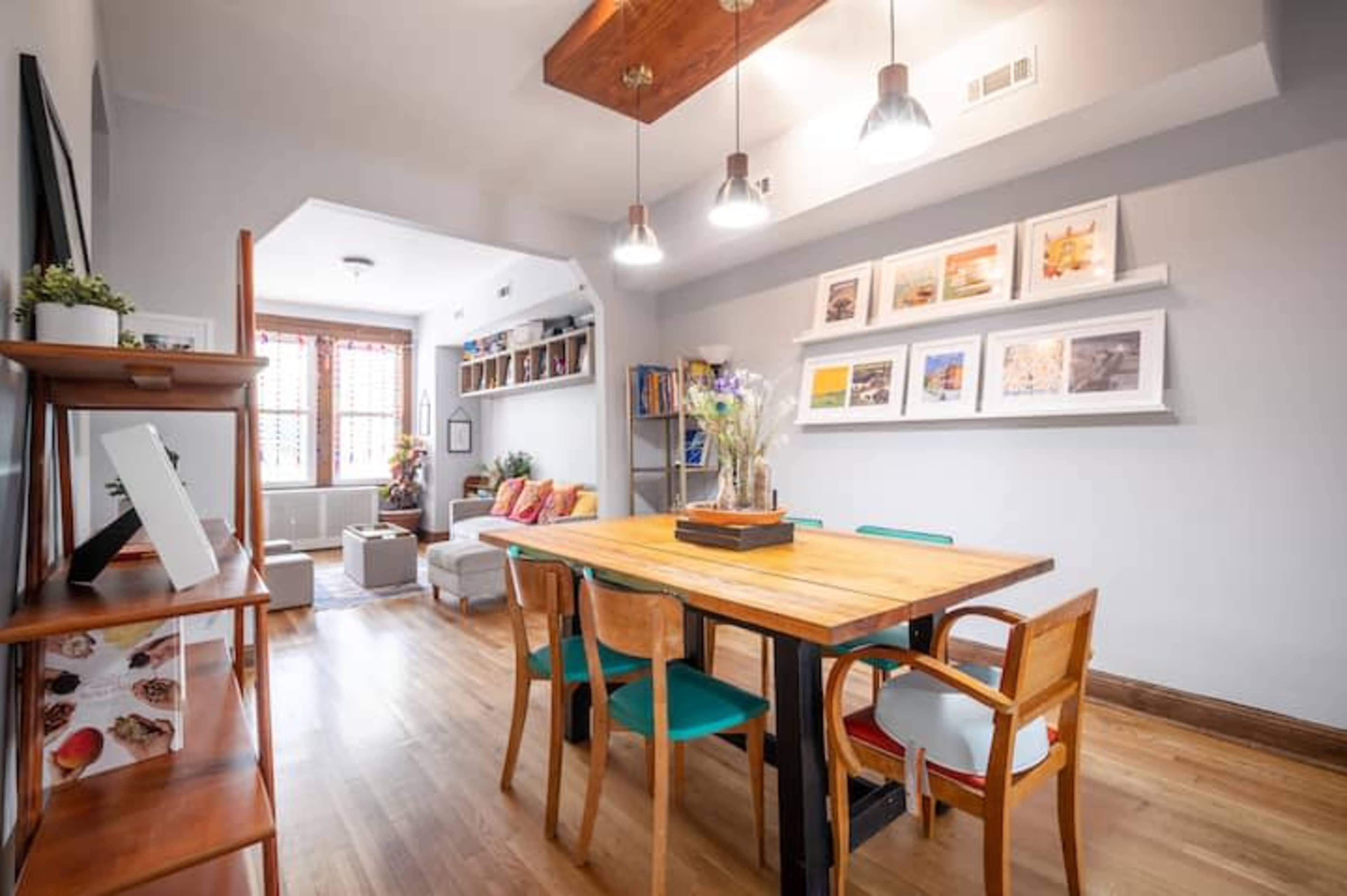 A dining area features a wooden table surrounded by colorful chairs, with framed photos on the wall and a shelf displaying books and decorative items.