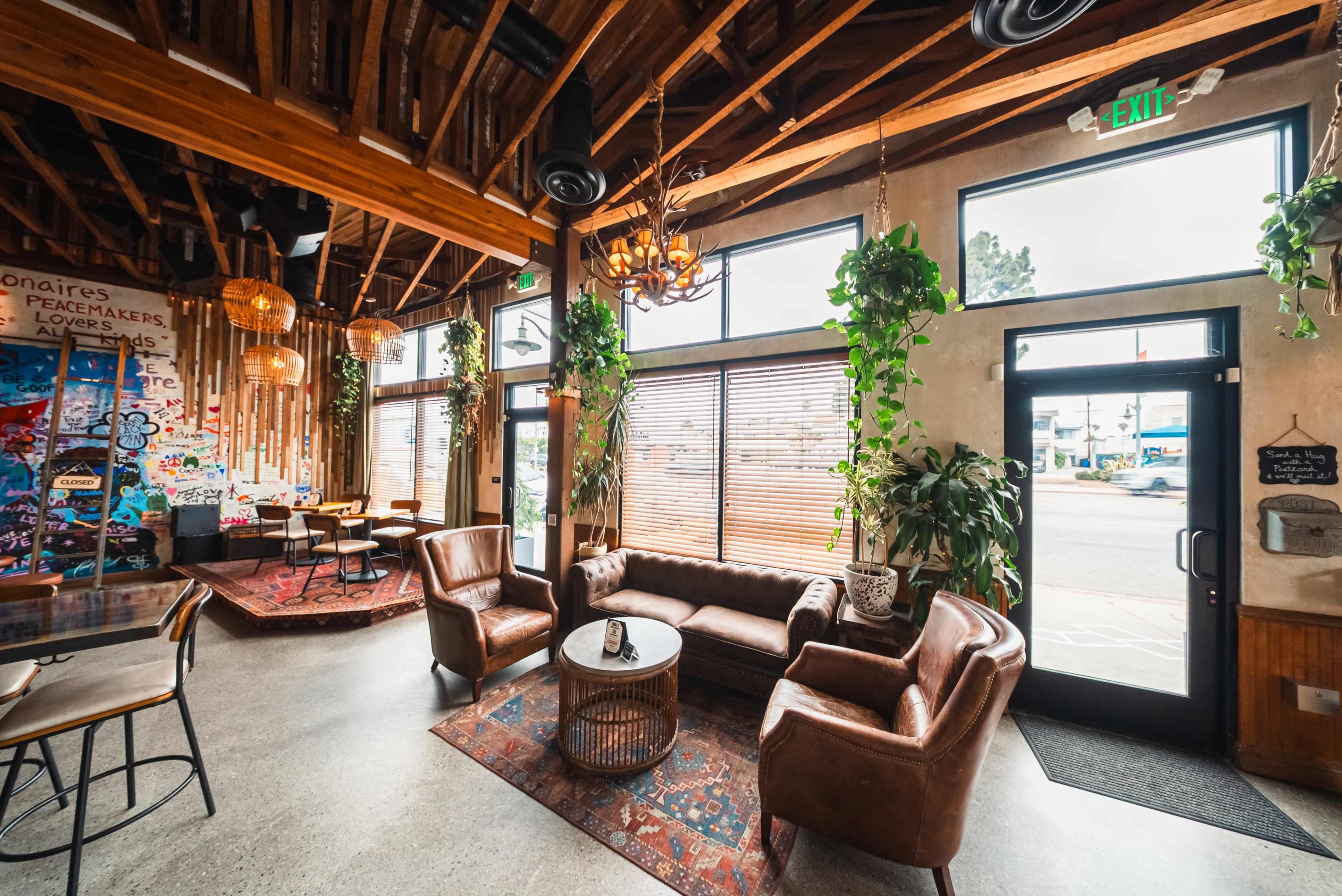 The image shows a stylish café interior with wooden beams, comfortable leather seating, and large windows adorned with plants.