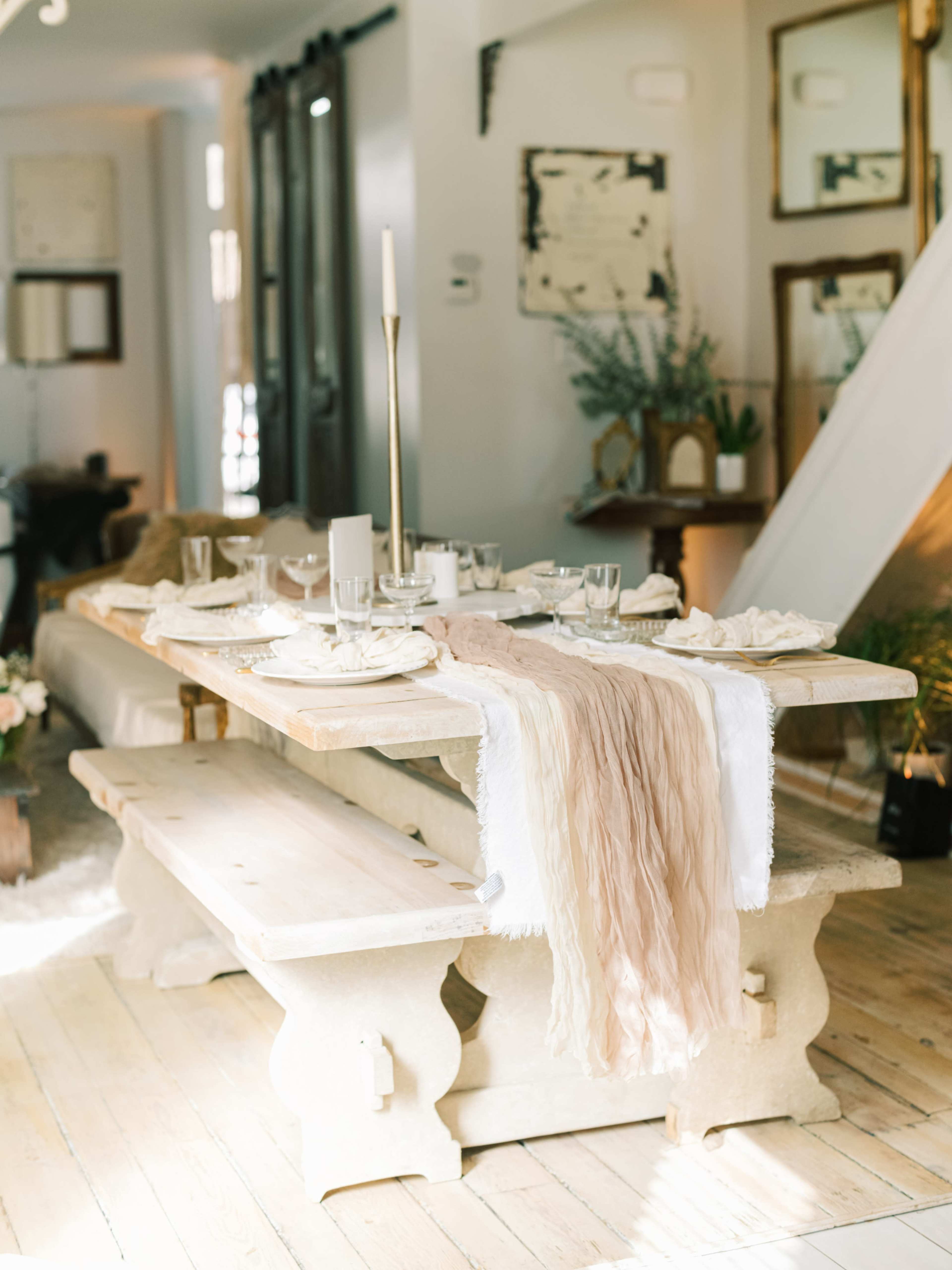 A rustic wooden dining table is set with glassware and a soft runner, surrounded by light-filled decor and mirrors.