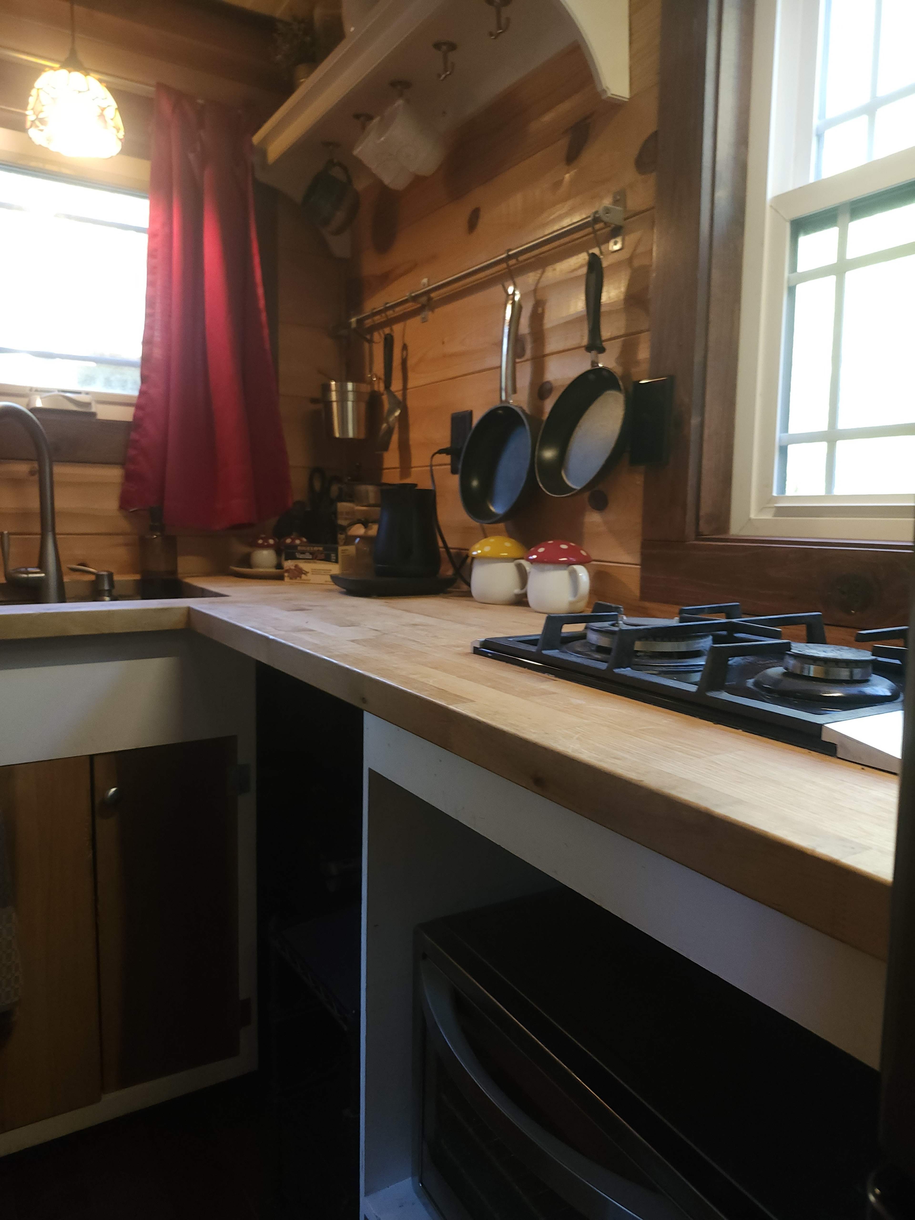 A small kitchen scene features a wooden counter, gas stove, hanging cookware, and red curtains by the window.
