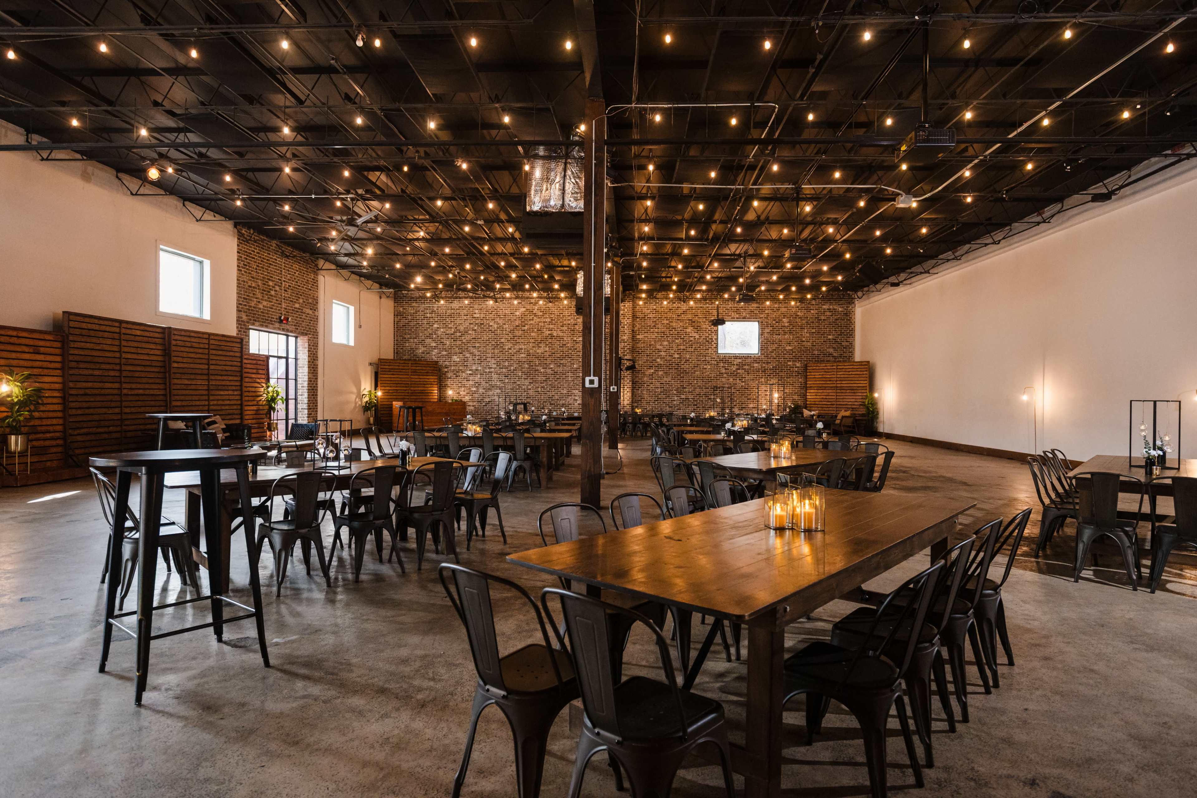 The image shows a spacious event venue with wooden tables and metal chairs, illuminated by overhead string lights, featuring brick walls and a concrete floor.
