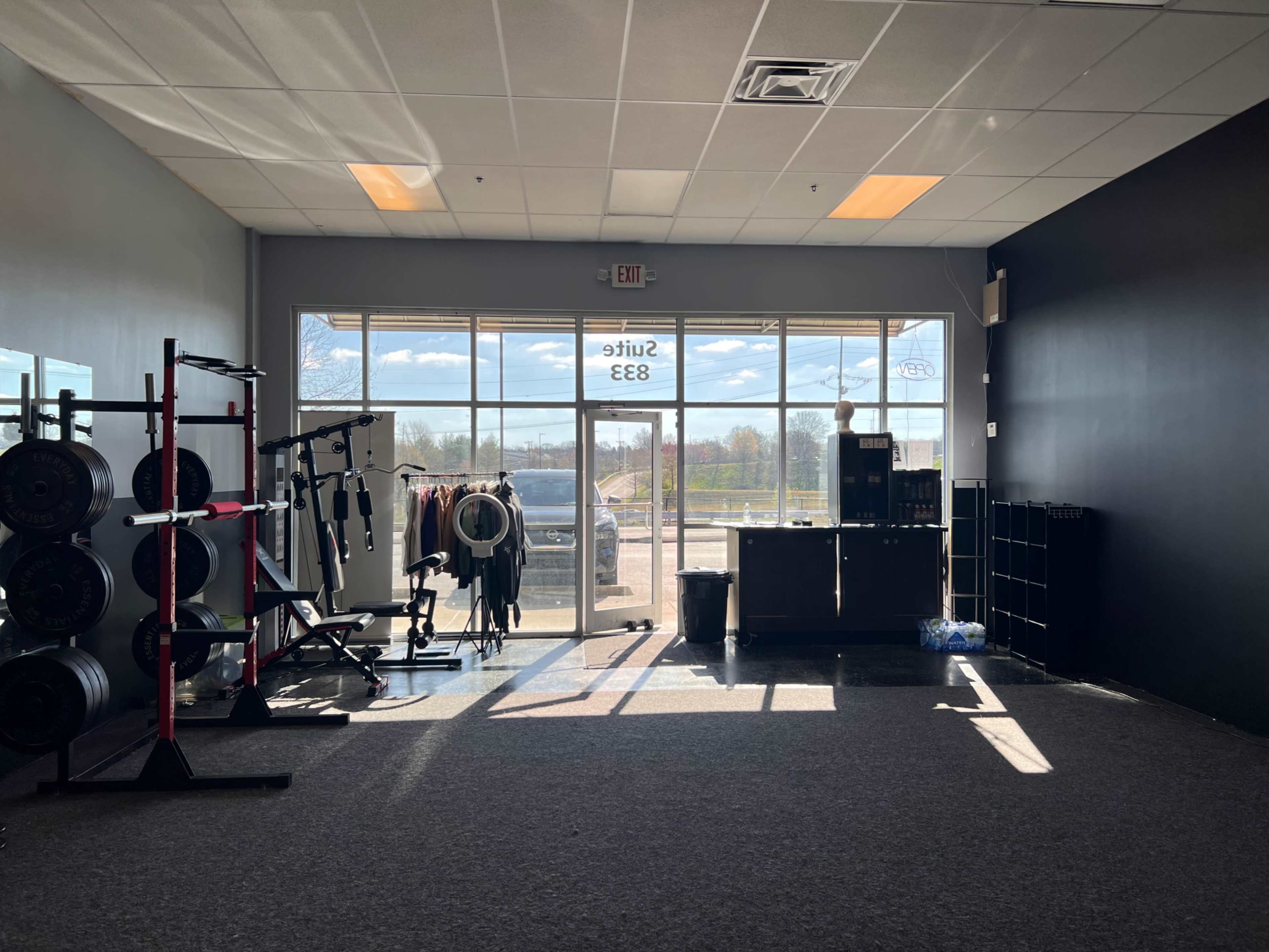 A fitness studio with weightlifting equipment and a glass door leading outside, illuminated by natural light.