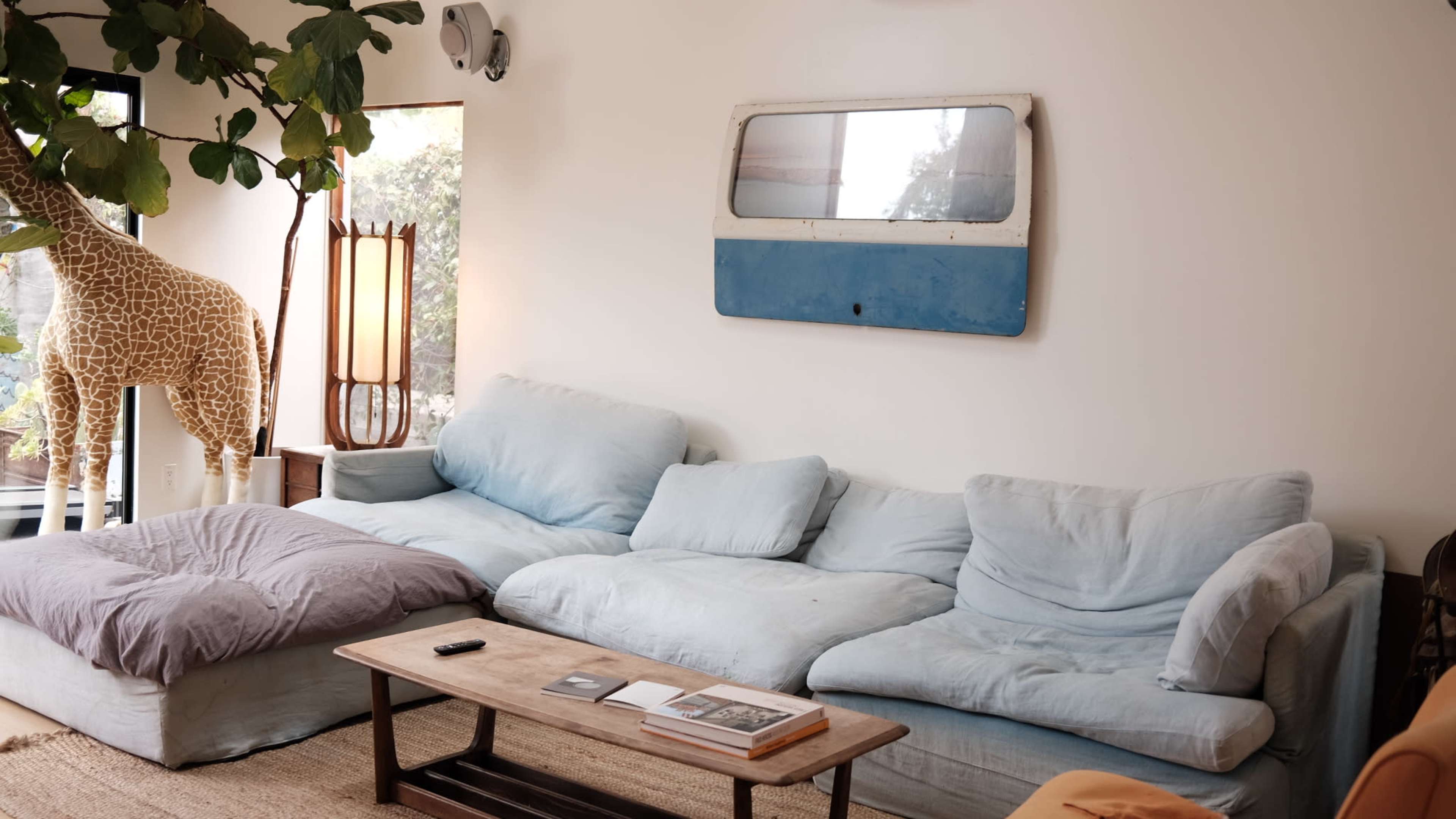 A living room features a large, soft blue couch, a wooden coffee table, and a giraffe statue beside a lamp and a window with natural light.