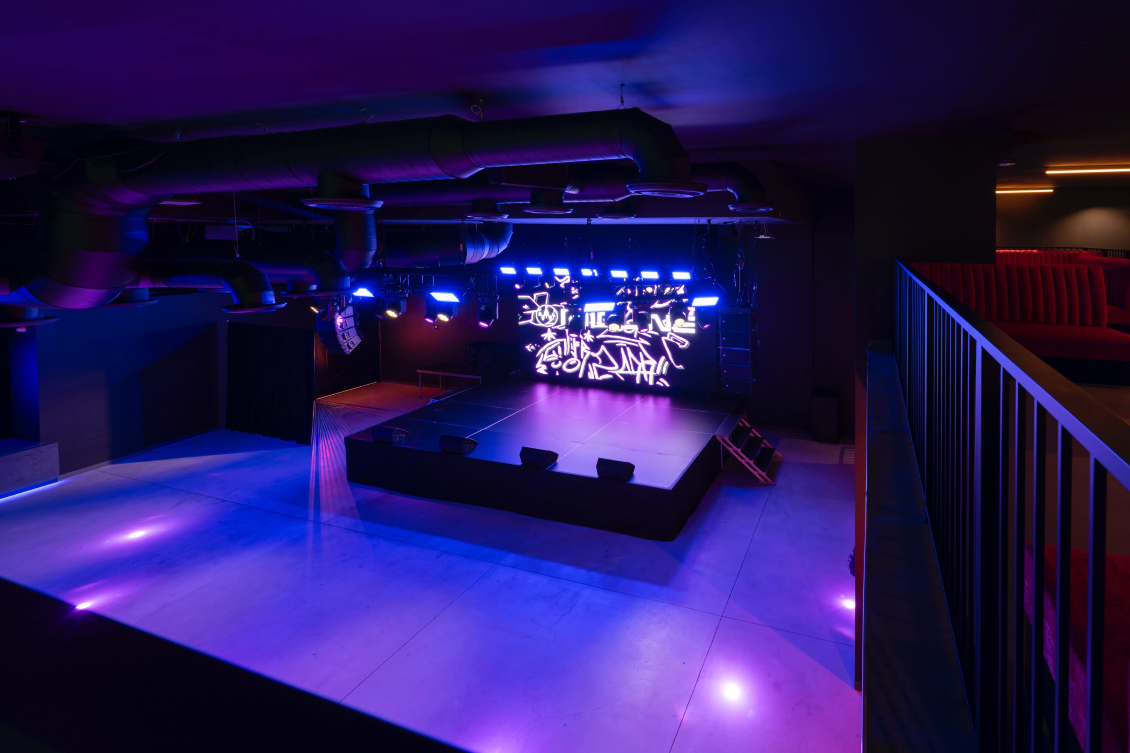 The image depicts an empty music venue featuring a raised stage illuminated by colorful LED lights and a digital backdrop.
