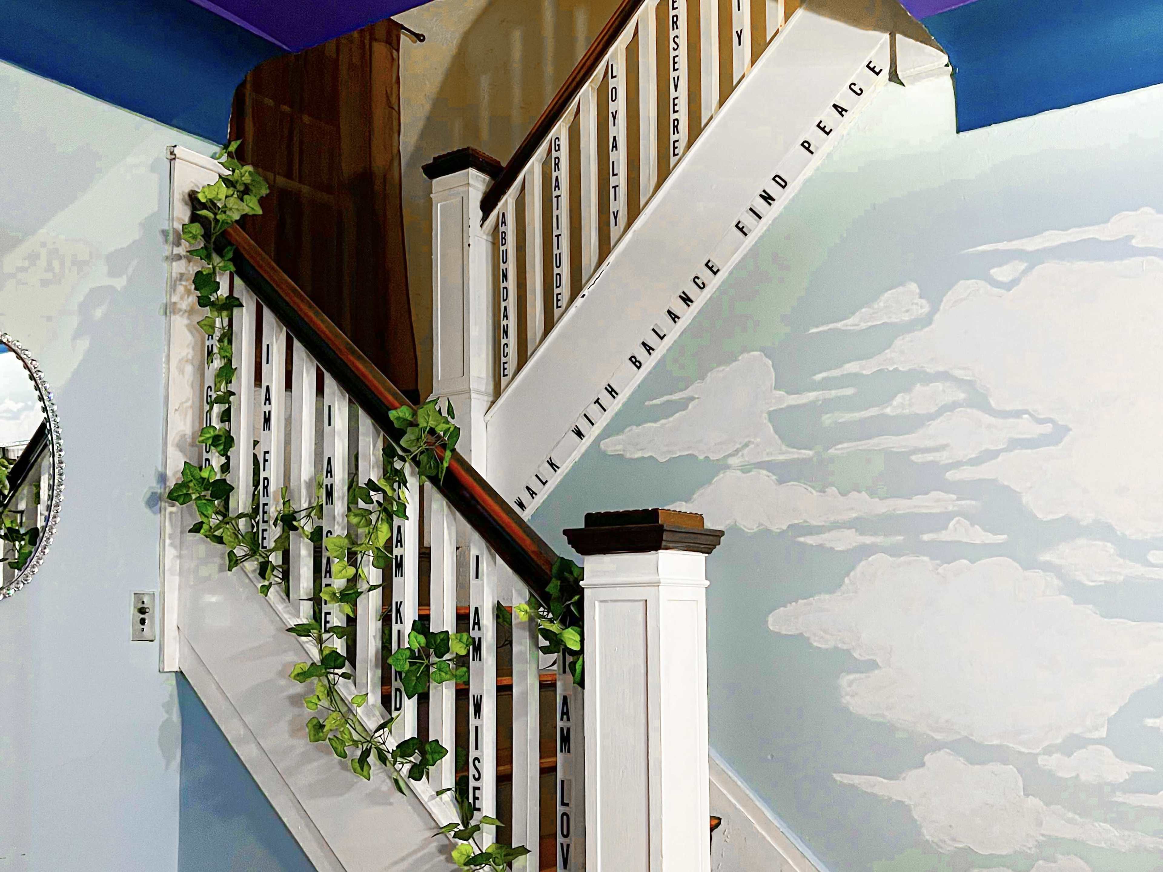 A staircase with a wooden railing adorned with green vines leads to a wall featuring painted clouds and inspirational phrases.