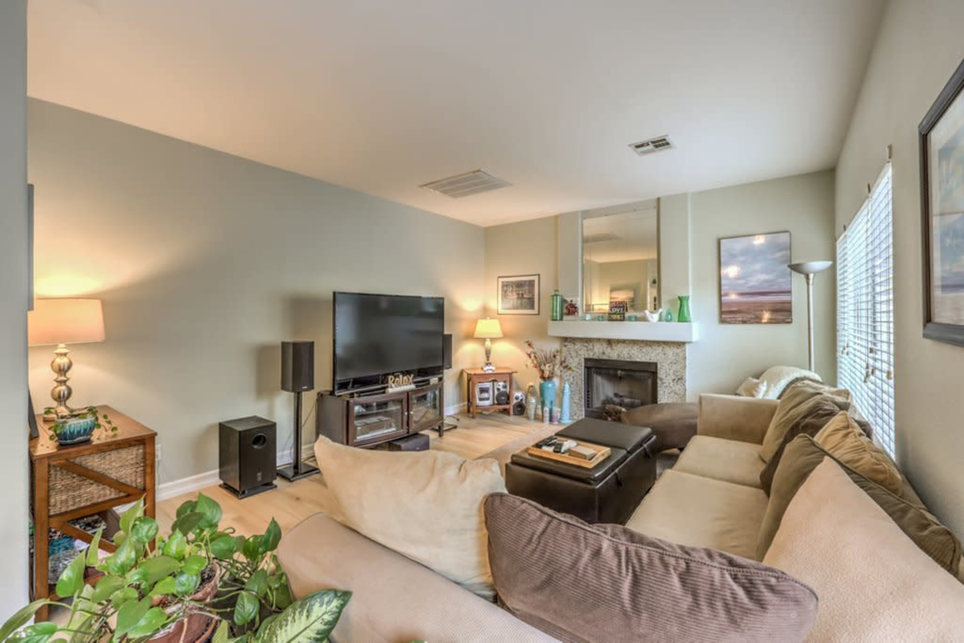A living room features a sectional sofa, a television stand, a fireplace, and decorative elements including plants and framed pictures.