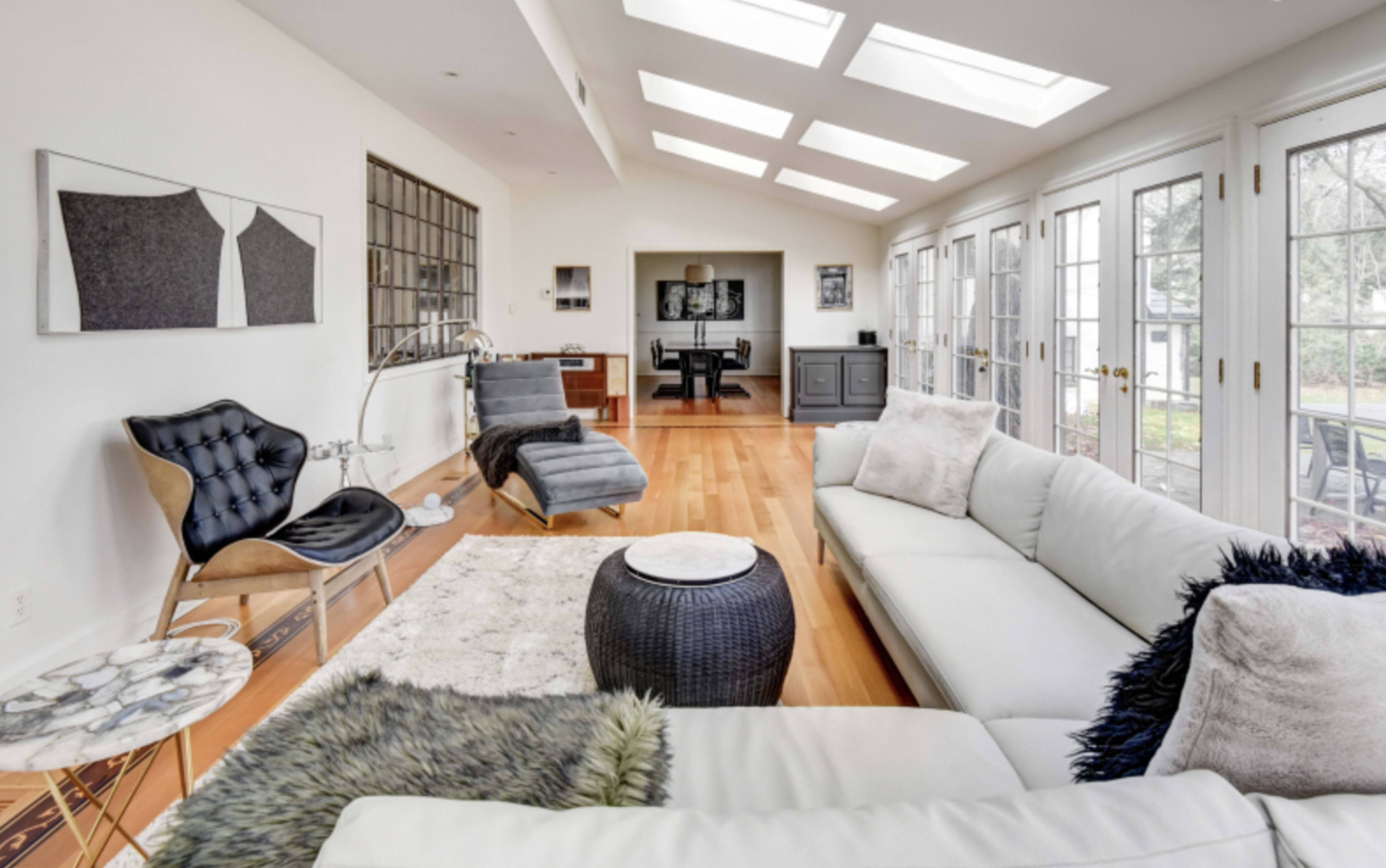 A modern living room with large windows, wooden flooring, and minimalist furniture including a white sectional sofa and a sleek chair.
