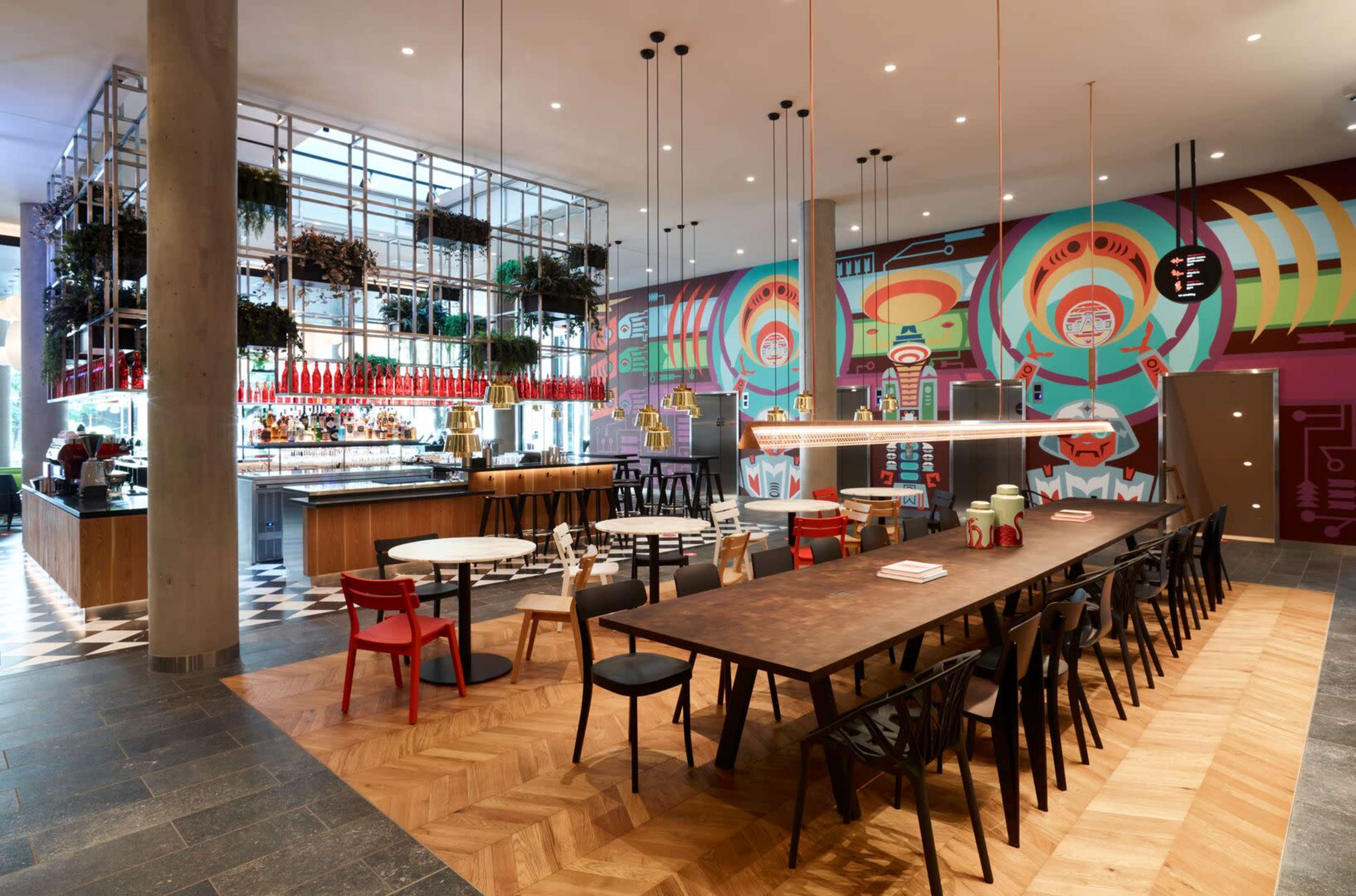 A modern restaurant interior featuring a large communal dining table, colorful murals, and a bar area with shelves of plants and bottles.