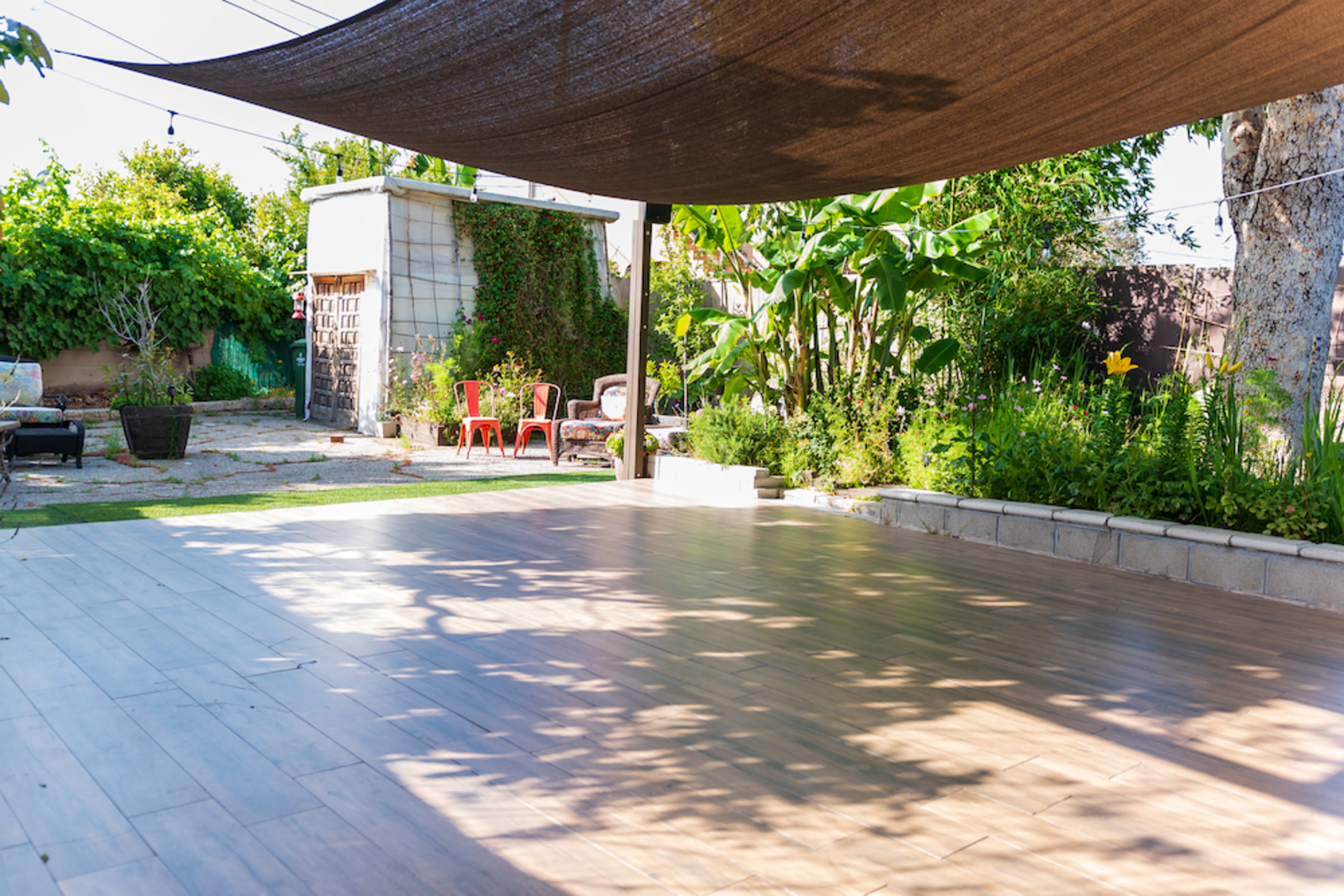 The image shows a patio with wooden flooring shaded by a sail canopy, surrounded by greenery and outdoor seating.