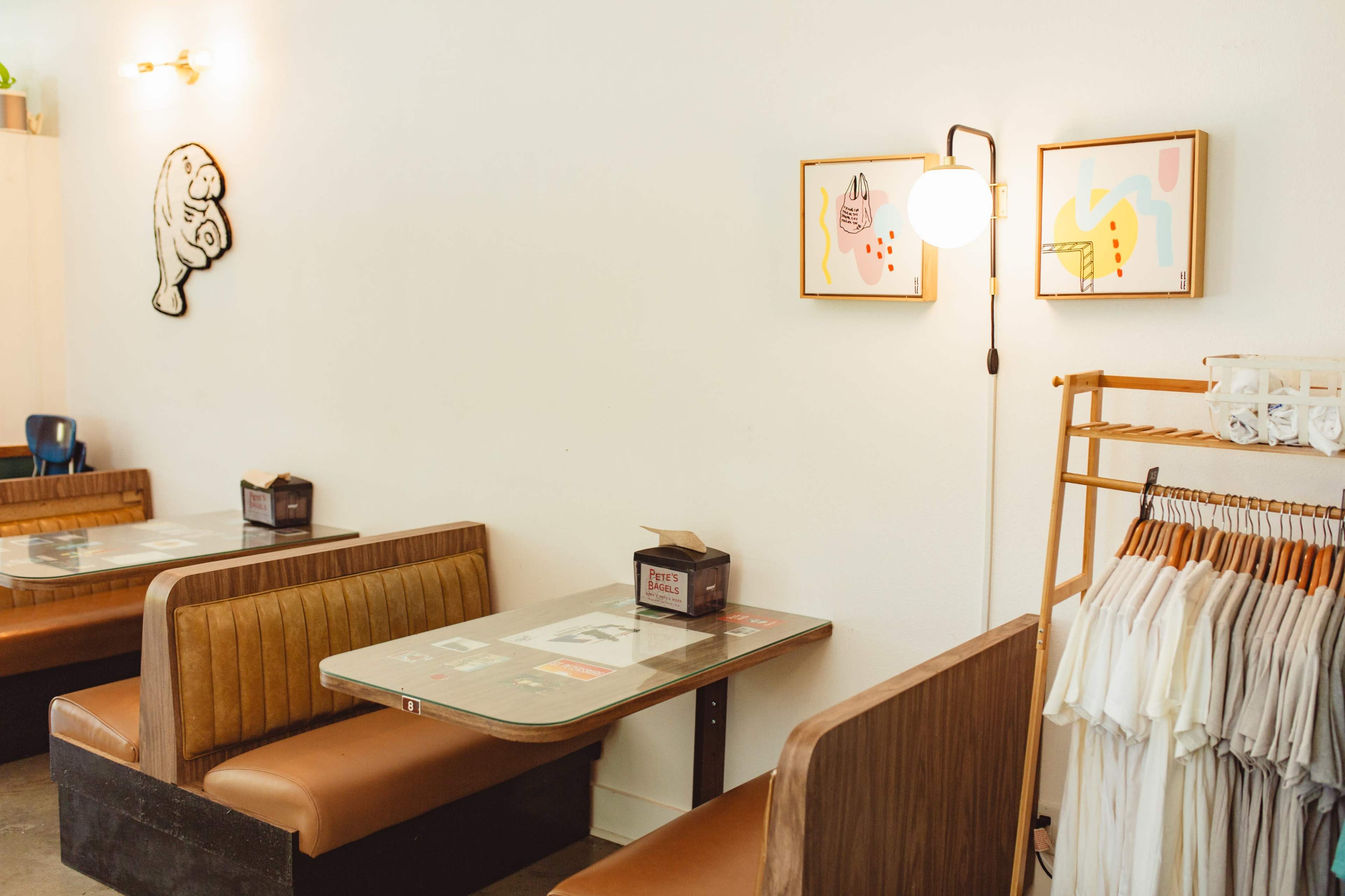 The image shows a cozy cafe interior with wooden seating, framed artwork on the walls, and a clothing rack displaying shirts.