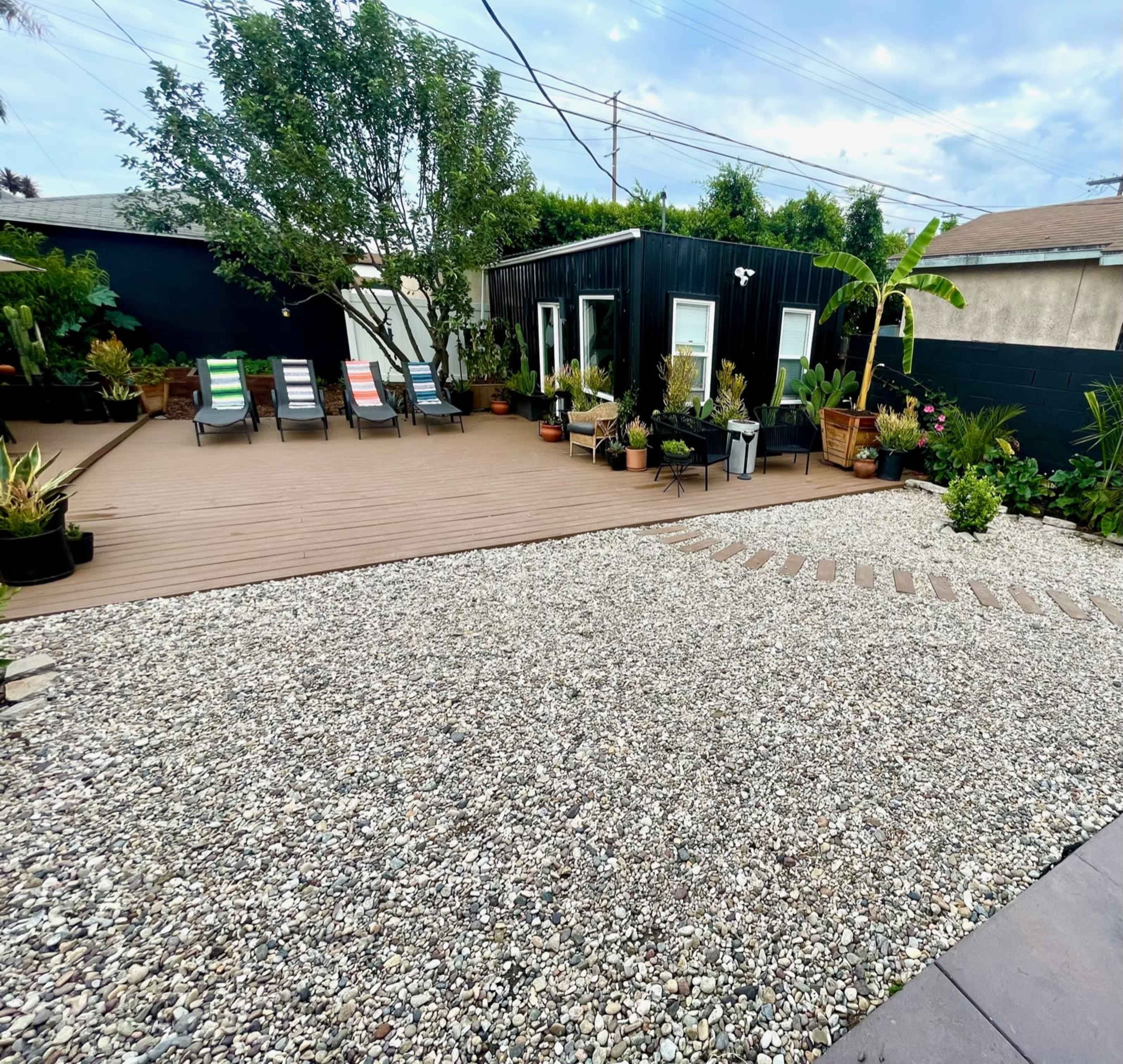 The image shows a landscaped backyard featuring a wooden deck with lounge chairs, gravel pathways, and various potted plants surrounding a dark-colored building.